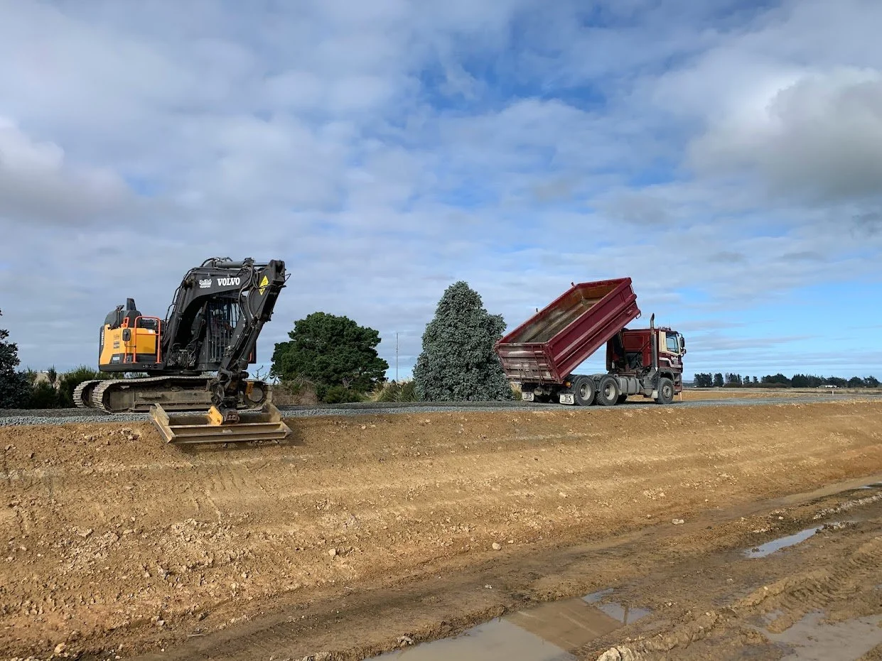 A construction site with a Volvo excavator and a red dump truck on a dirt path under a partly cloudy sky.
