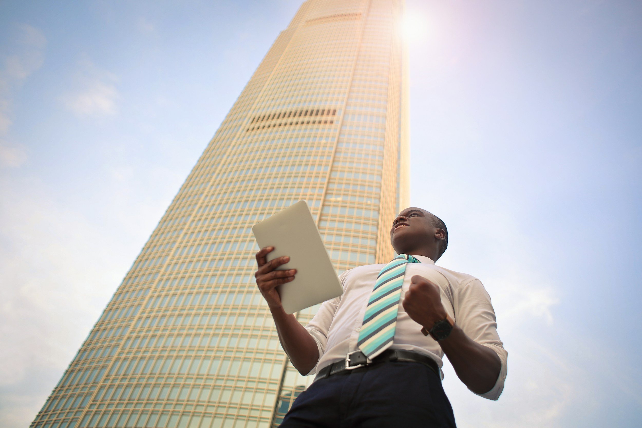 An upward low-angle view of a young man in business attire holding a tablet, with a tall skyscraper and blue sky in the background.