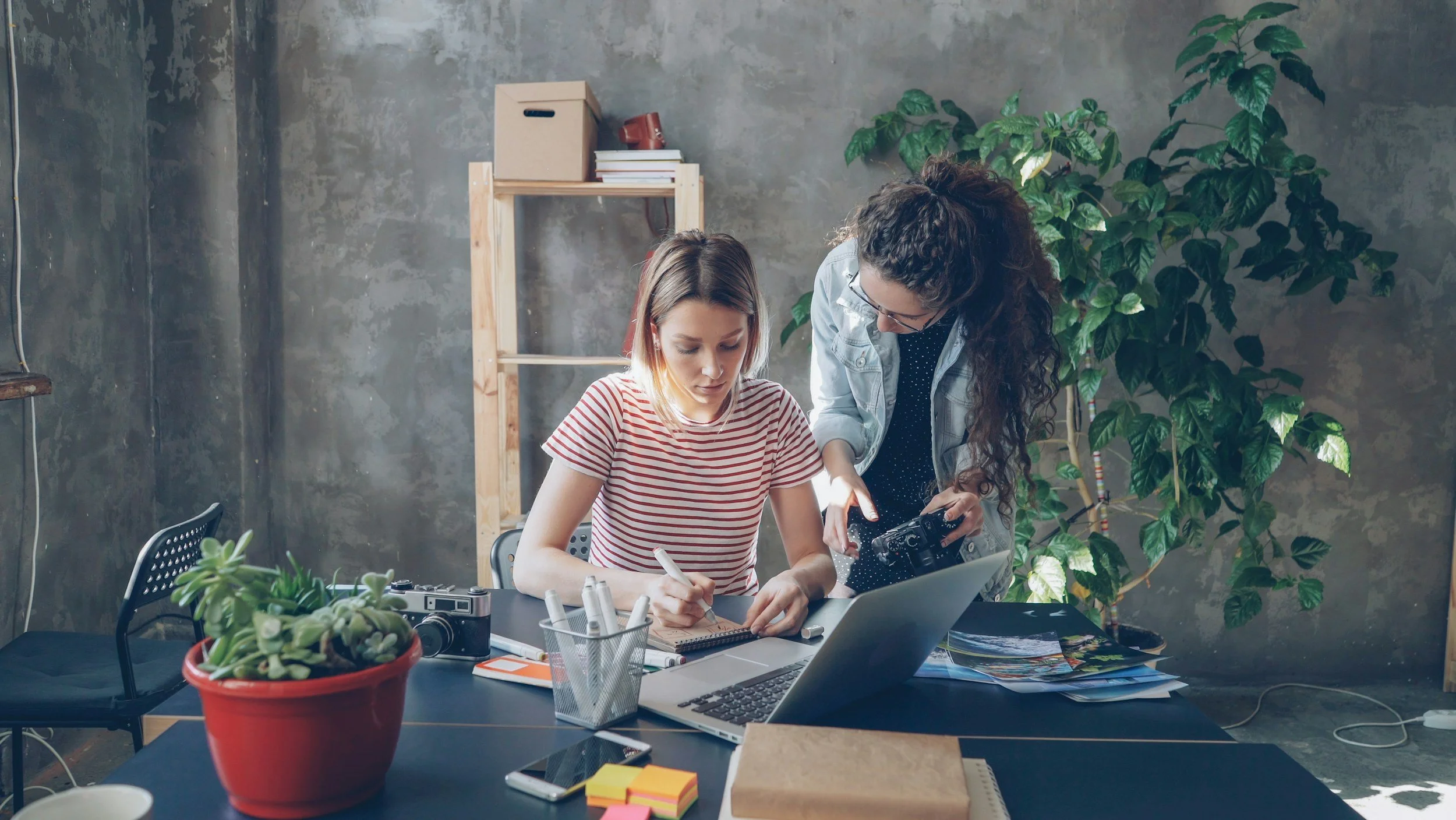 Two women working together at a desk with plants and office supplies in an industrial-style room.