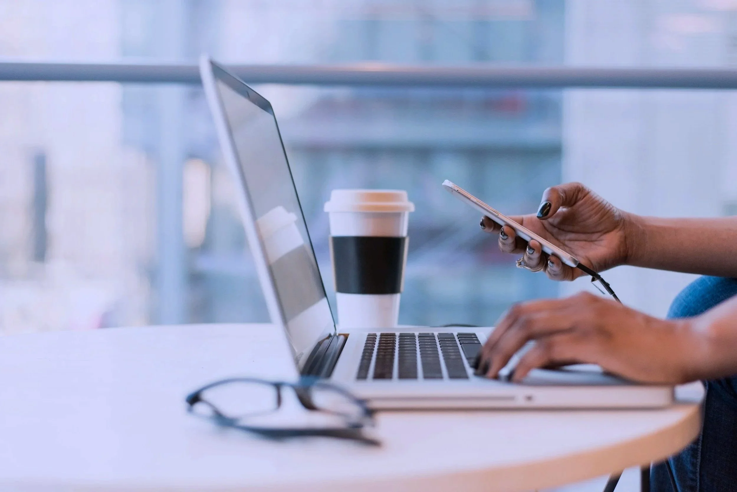 Person using a smartphone at a work desk with a laptop and coffee cup.