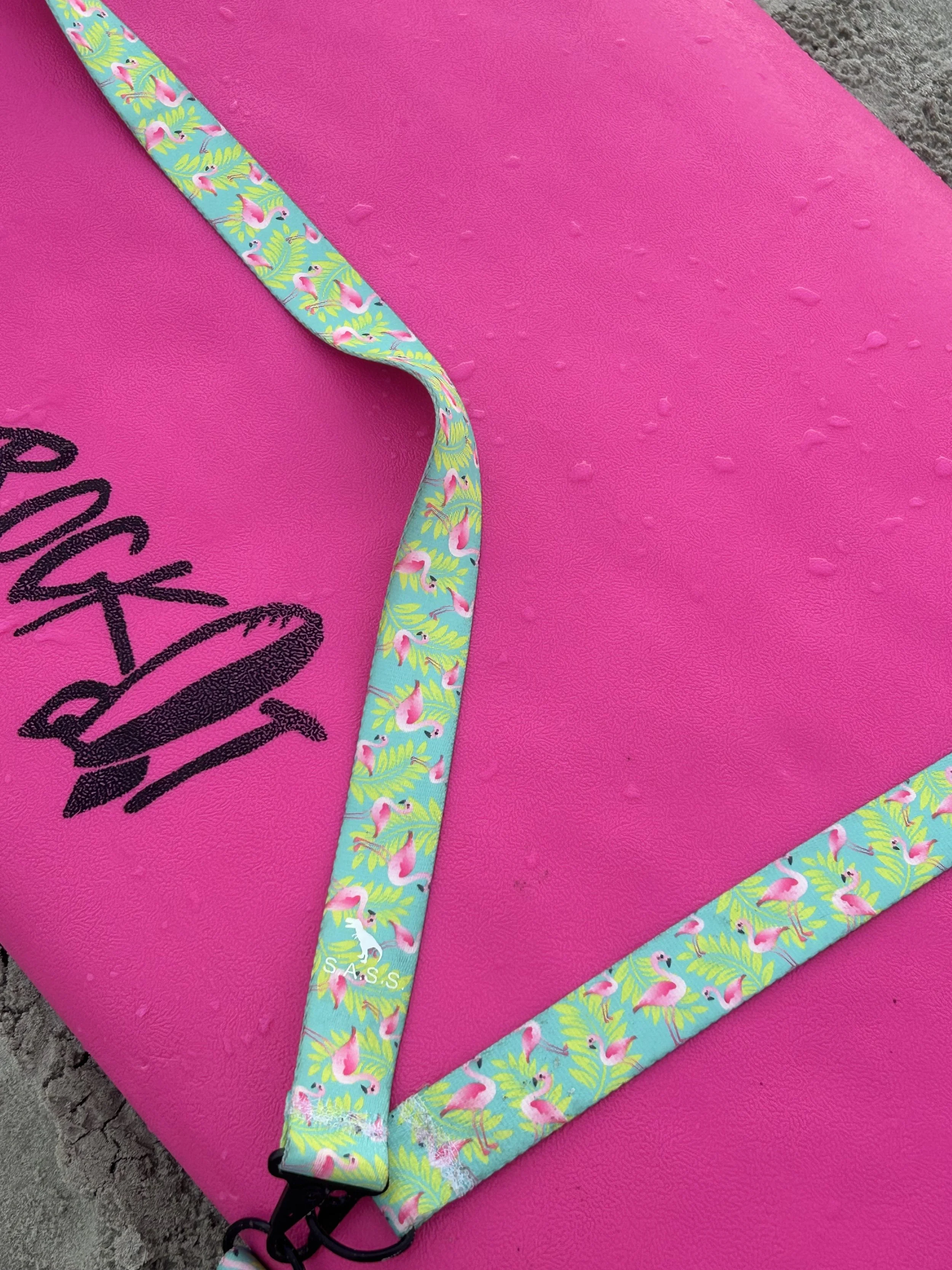 Close-up of a pink paddleboard with black logo of a hat and sunglasses, decorated with a colorful strap featuring pink flamingos and green foliage, positioned on a sandy surface.
