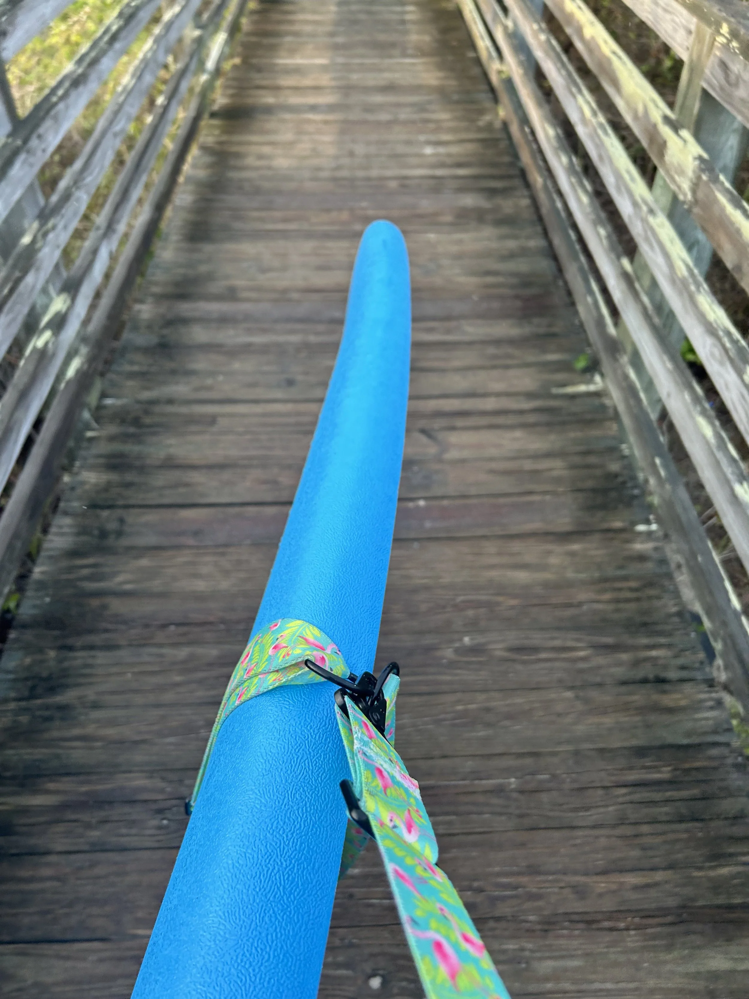 View from above of a blue foam surfboard with a colorful strap, on a wooden bridge.