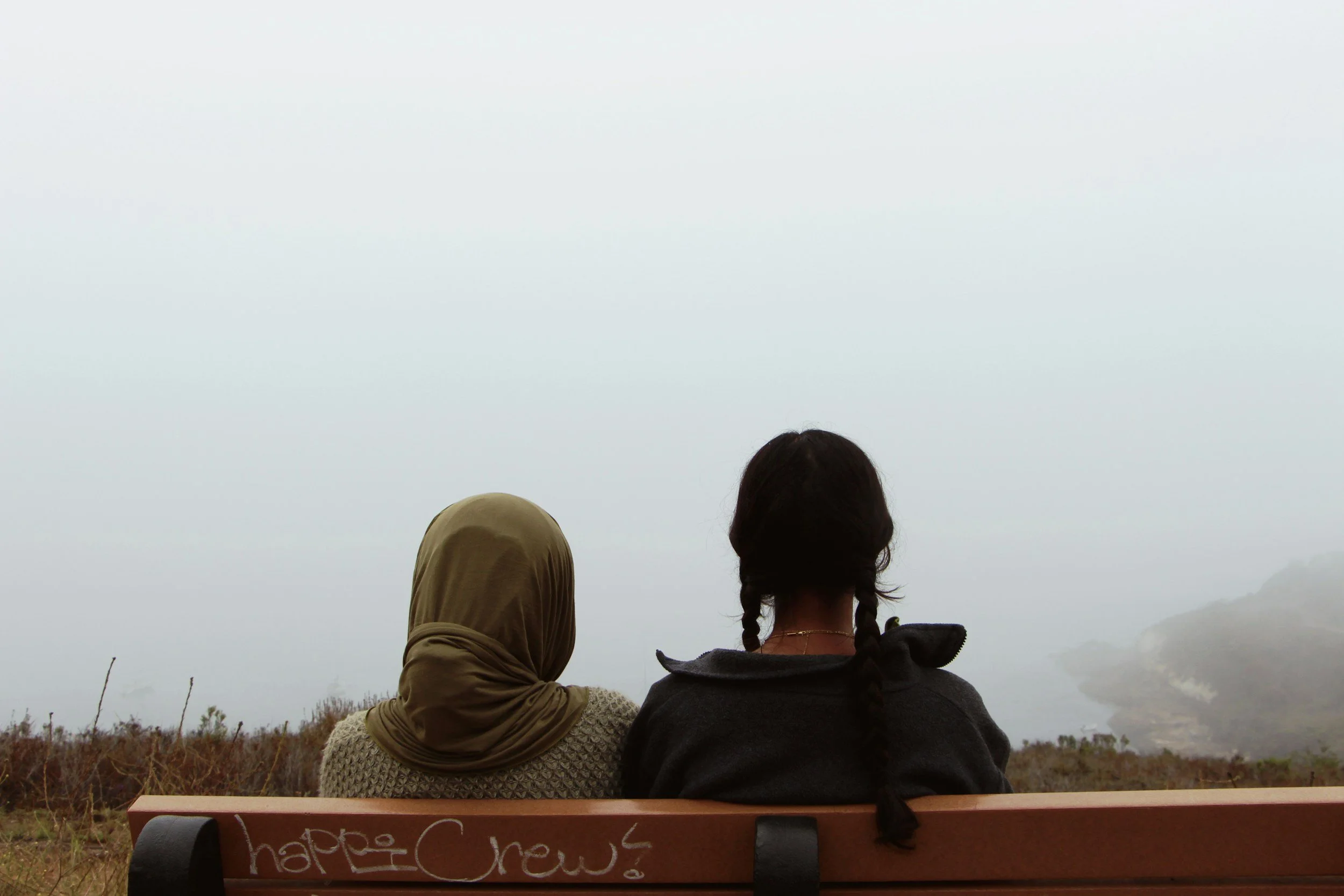 Two women sitting on a bench outdoors, facing away, one wearing a headscarf and the other with braided hair, near a foggy coastline.