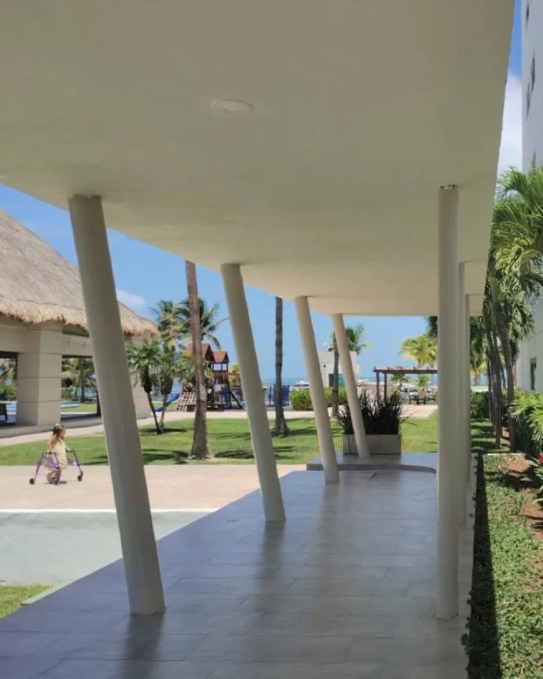 Vista desde un pasillo cubierto con columnas inclinadas, en un entorno tropical con palmeras, césped, y una niña jugando en el parque.