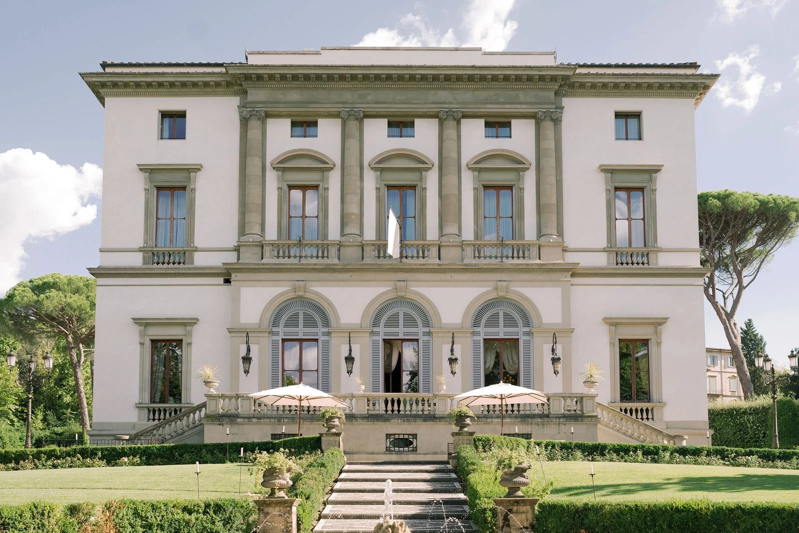 A luxurious three-story mansion with classic architecture, large windows, and ornate details. The front yard has a lawn, steps leading up to the entrance, and two umbrellas with chairs underneath. There are trees and greenery surrounding the house.