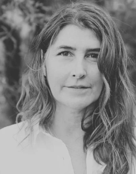 Black and white portrait of a woman with wavy hair, looking at the camera with a slight smile, outdoors background