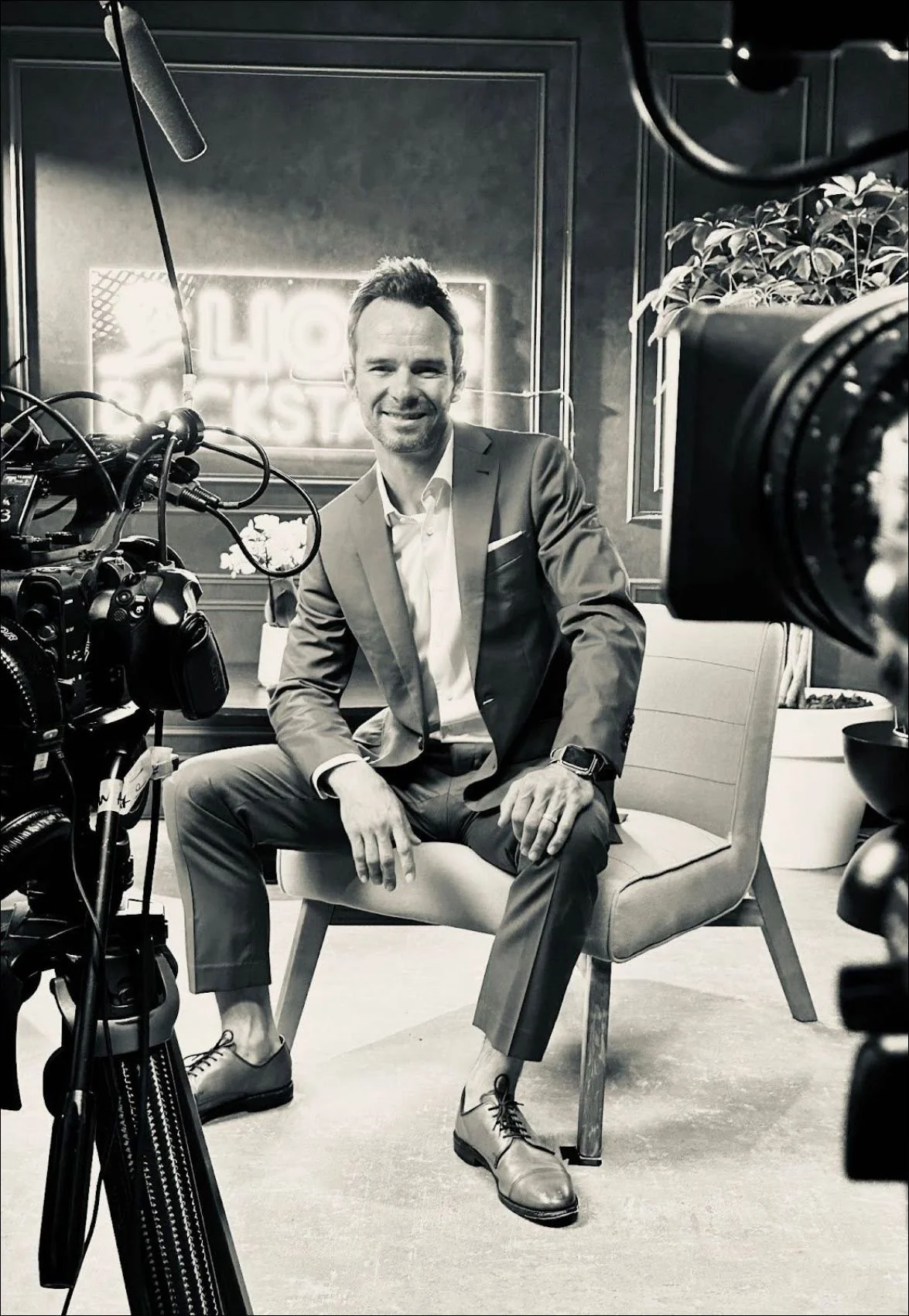 A man in a suit sitting on a chair during a video recording, surrounded by cameras and lighting equipment, with a neon sign and plants in the background.
