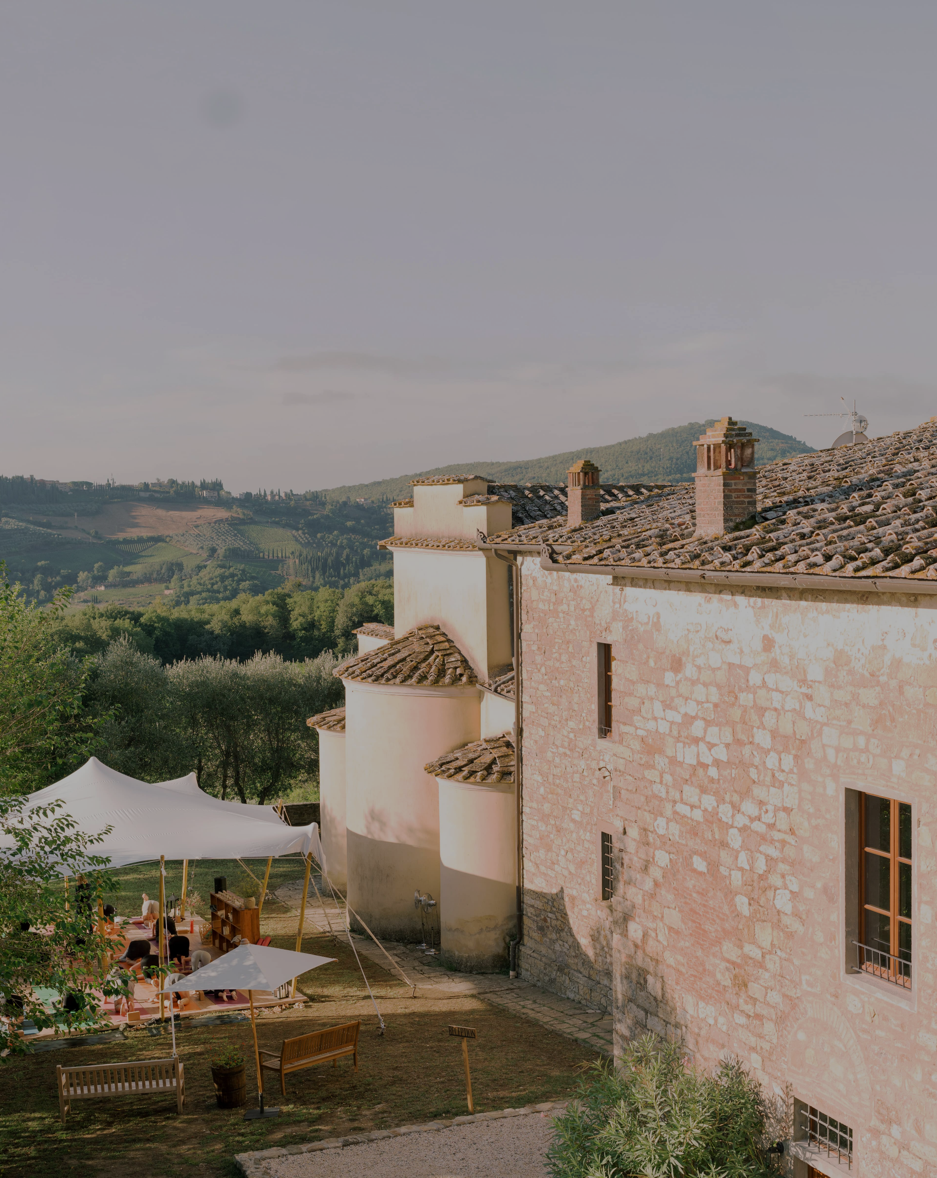 Rooftop view of an Italian countryside building with hills and vineyards in the background, and a tent with outdoor seating in the foreground.