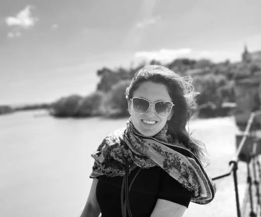 Woman wearing sunglasses and a scarf, smiling outdoors near a body of water with a castle or building on a hill in the background.