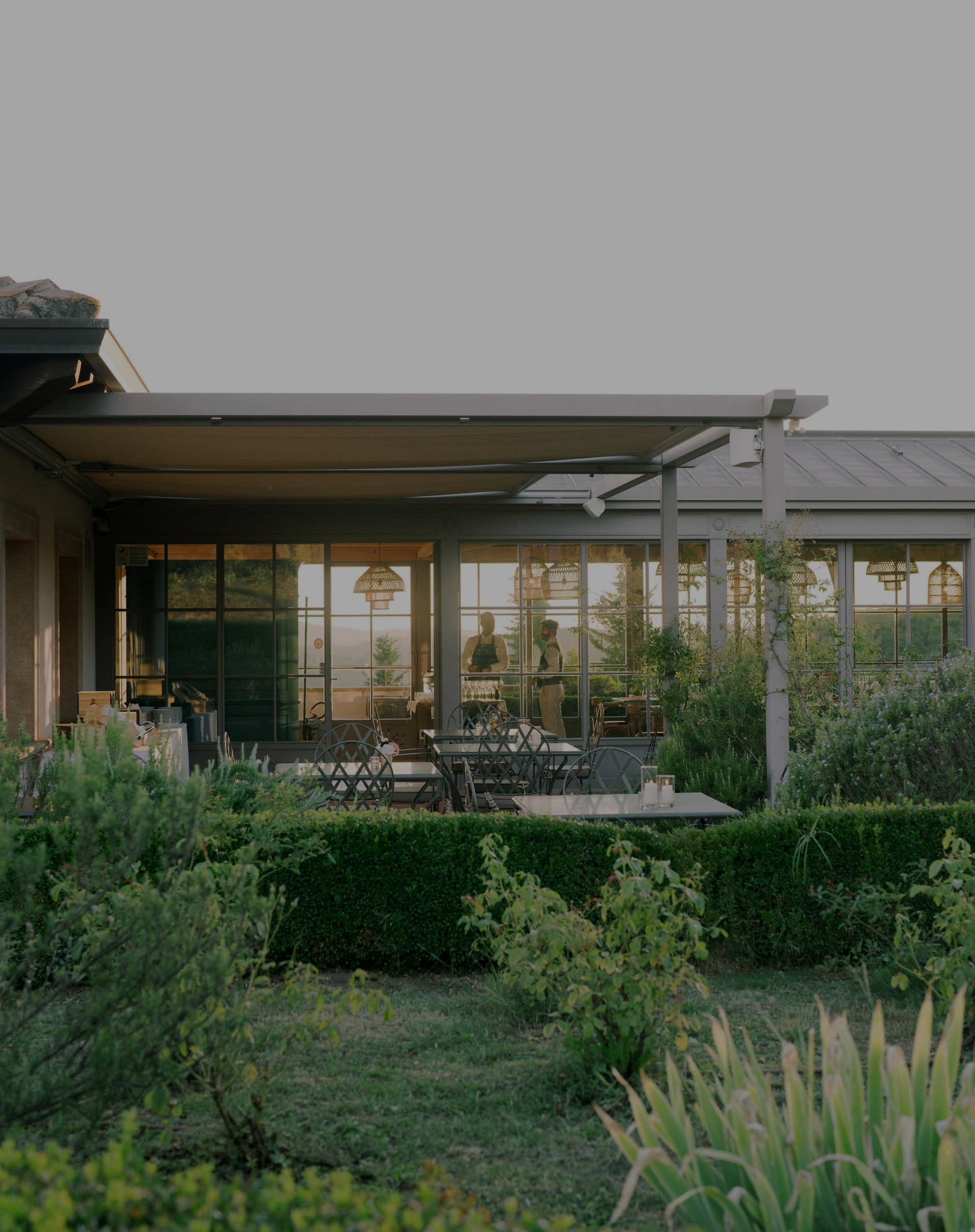 Exterior view of a restaurant patio with tables and chairs, greenery, and large windows showing two people inside at sunset.