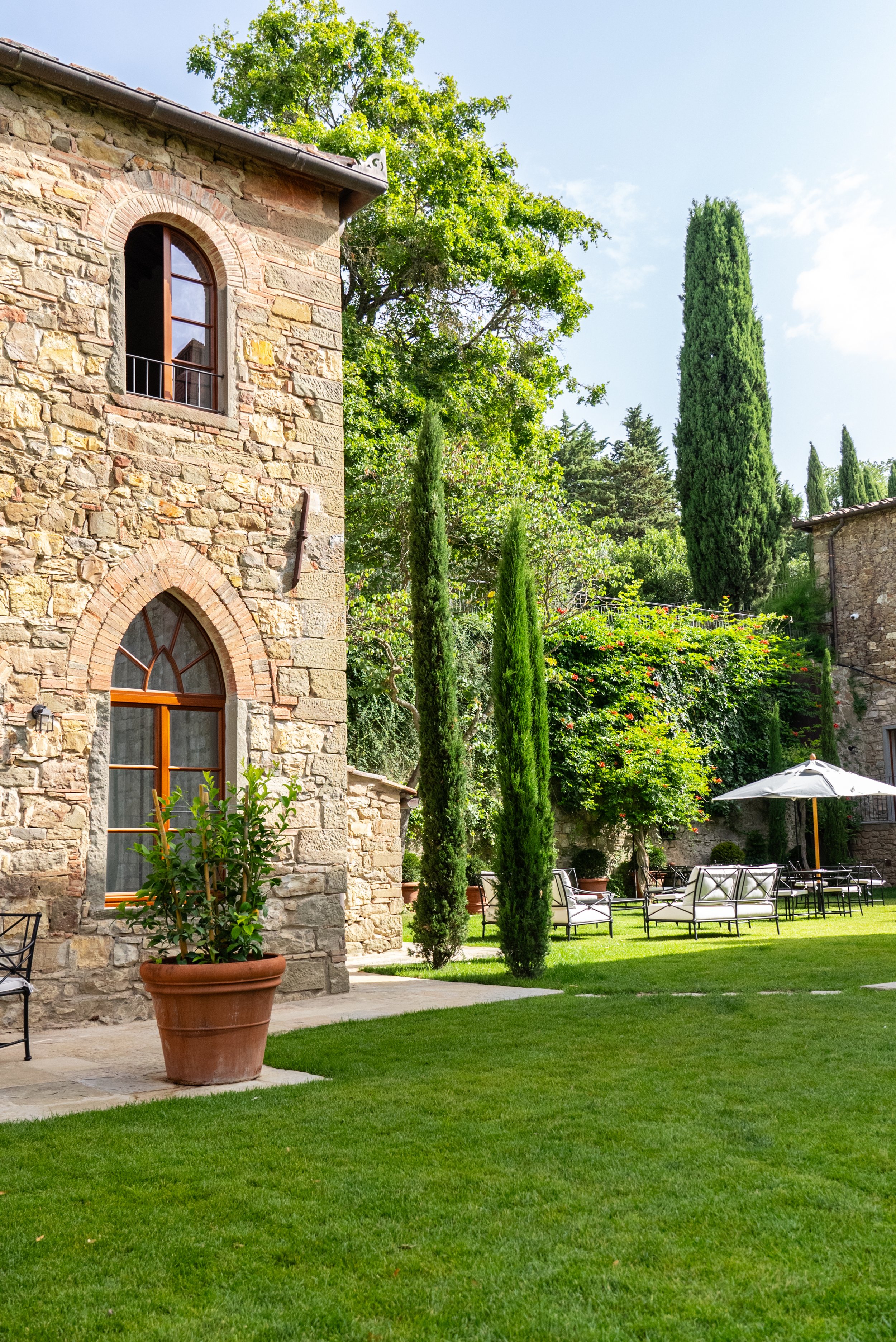A stone house with arched windows, surrounded by tall cypress trees, green lawn, outdoor furniture, and an umbrella in a lush garden under a clear sky.