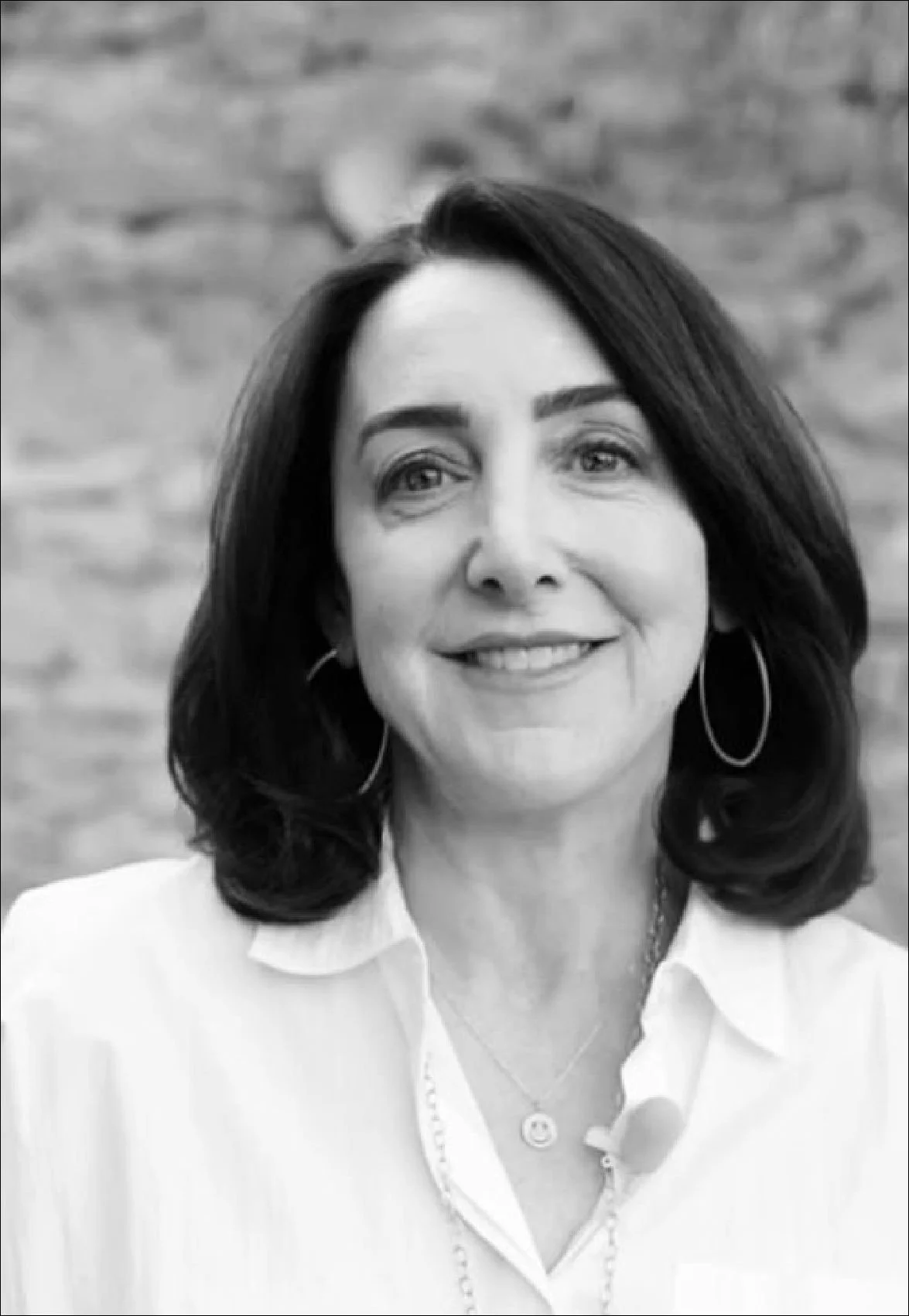 Black and white portrait of a woman with shoulder-length dark hair, hoop earrings, wearing a white shirt, smiling, in front of a blurred brick wall background.