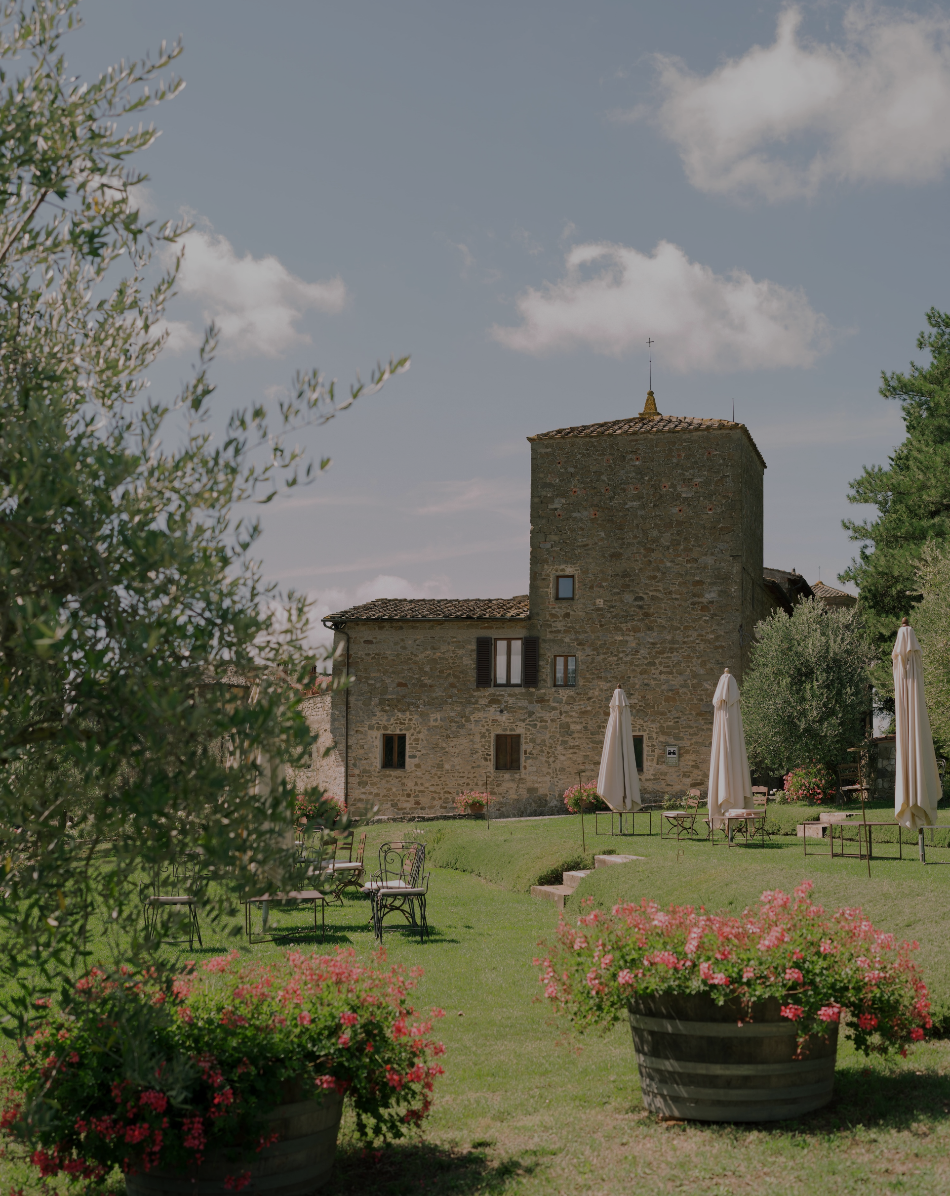 A stone building with a tower in a garden with tables, chairs, umbrellas, potted pink flowers, and green trees under a partly cloudy sky.