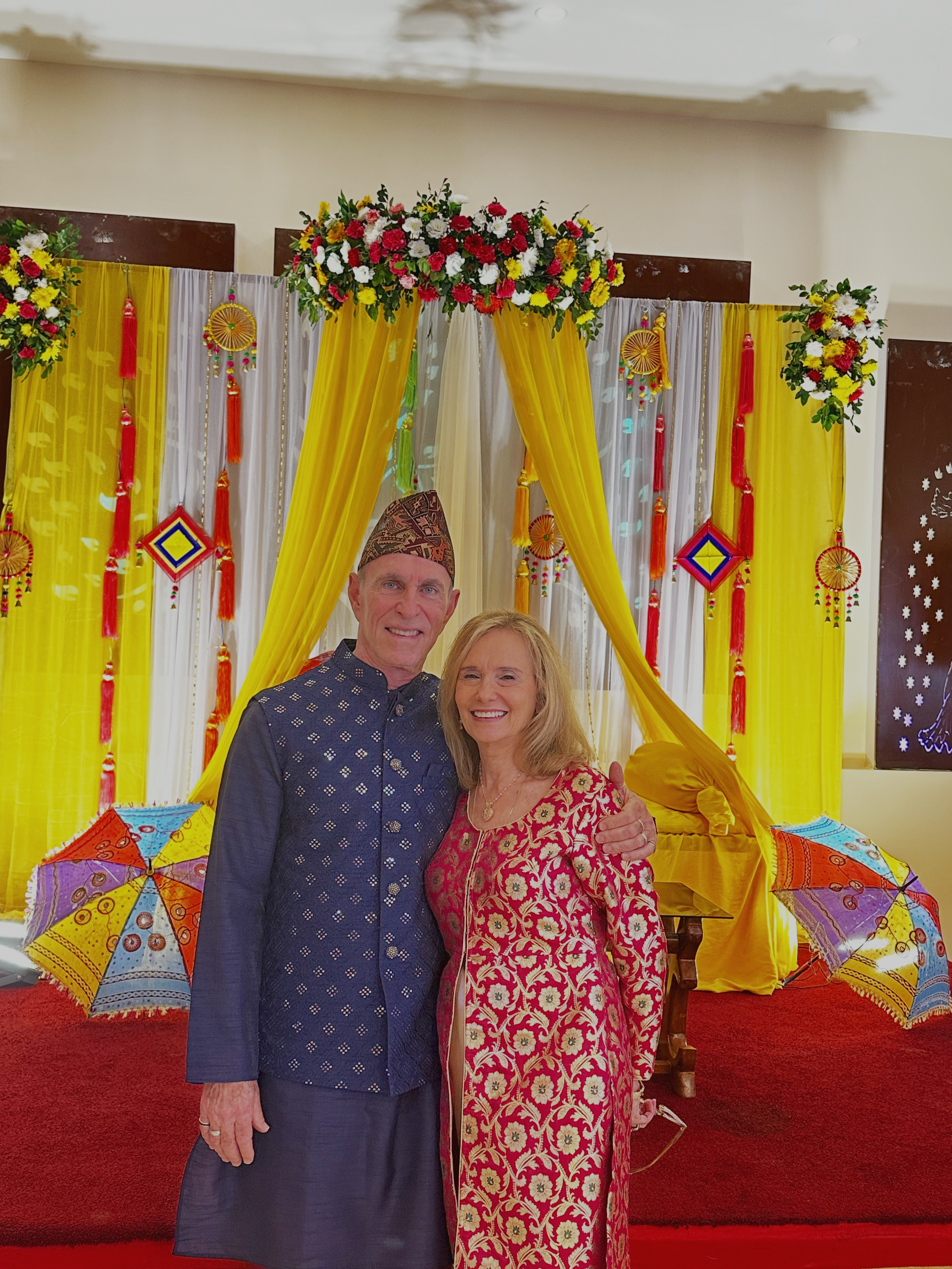 A man and woman dressed in traditional South Asian attire posing in front of a colorful decorated backdrop with yellow and white fabrics, flowers, umbrellas, and hanging ornaments.