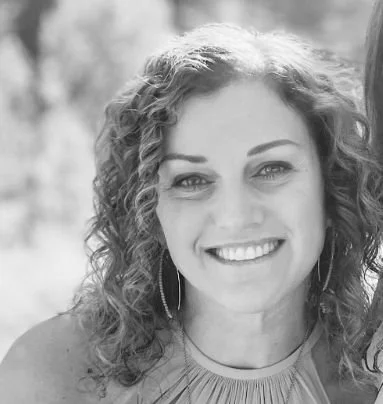 A woman with curly hair smiling outdoors in black and white.