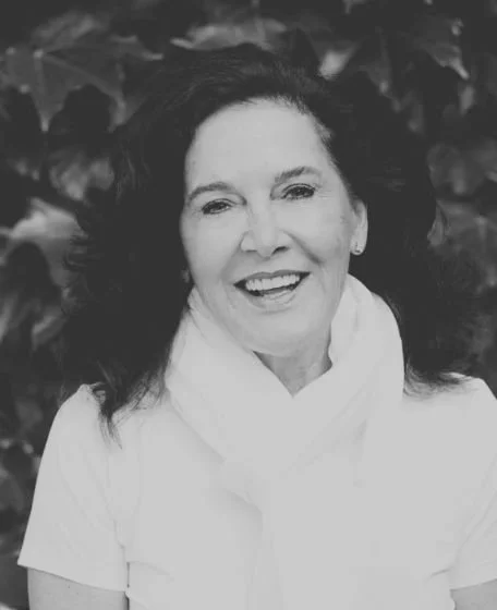 A smiling woman with dark, wavy hair in front of leafy background.