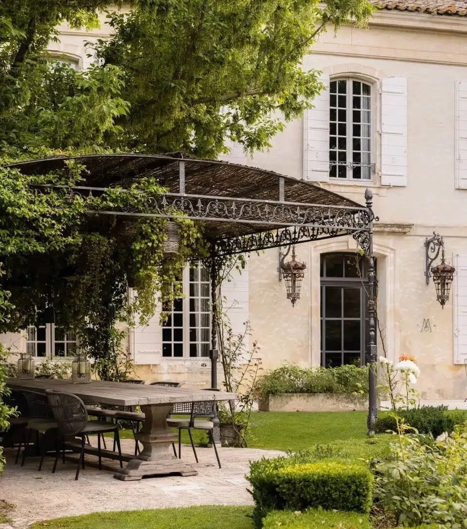 An outdoor patio with a dining table and chairs, surrounded by a garden with green shrubs and flowers, in front of a white house with tall windows and decorative shutters.