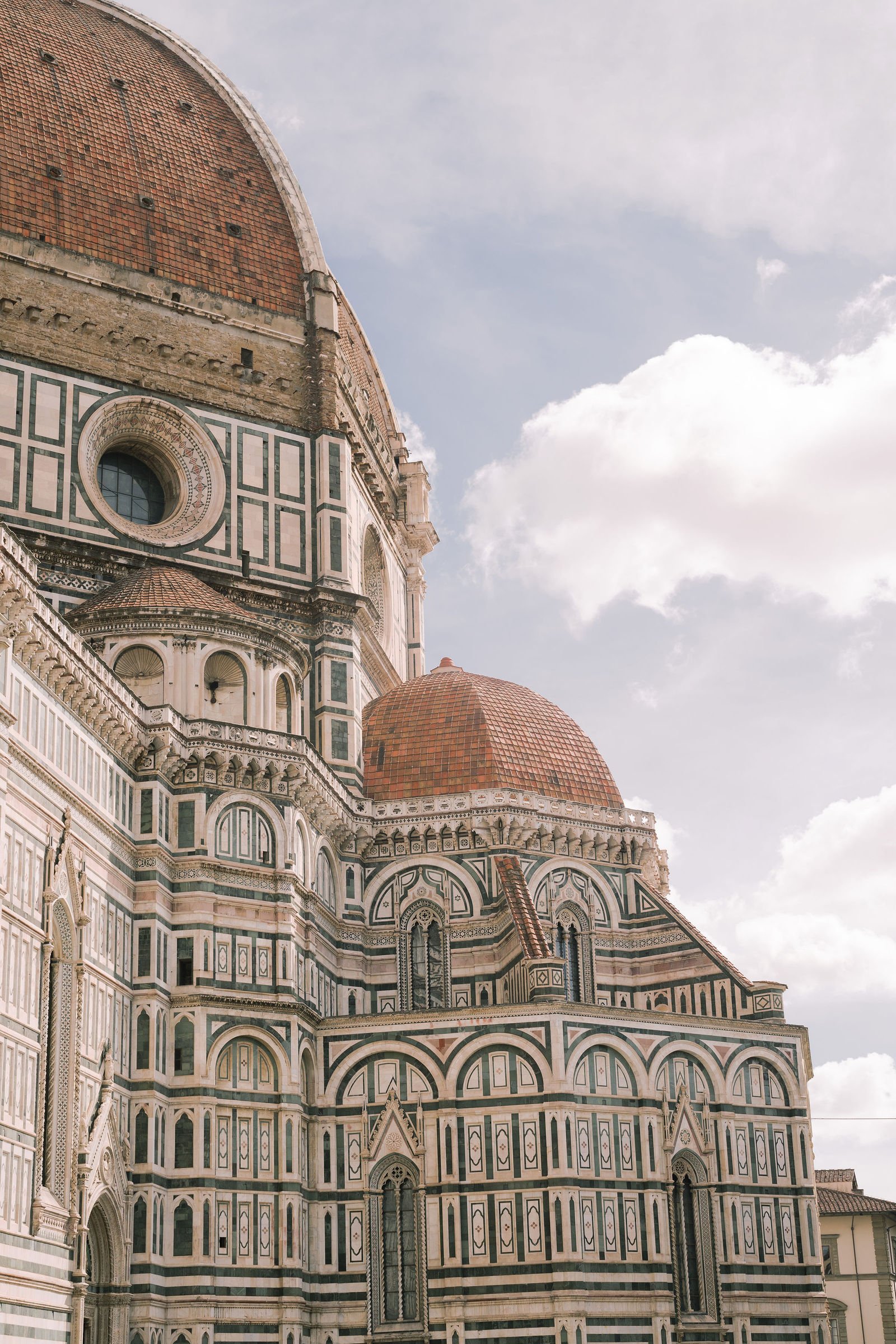 Close-up of the ornate exterior of the Florence Cathedral, also known as the Duomo, with detailed white, green, and pink marble patterns and a large dome with a reddish-brown tiled roof.