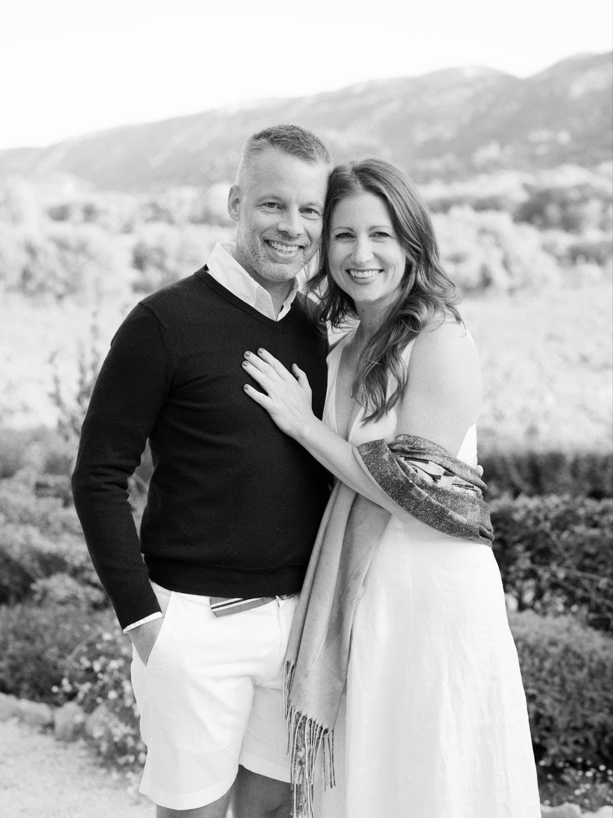 A couple smiling outdoors, dressed in casual and semi-formal clothing, standing close with a scenic background of mountains and greenery.