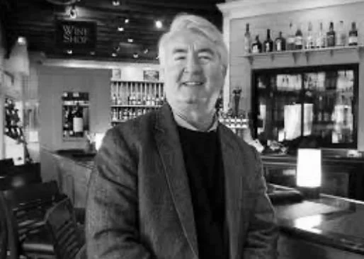 A smiling man with gray hair standing in a bar or restaurant with bottles on shelves behind him.