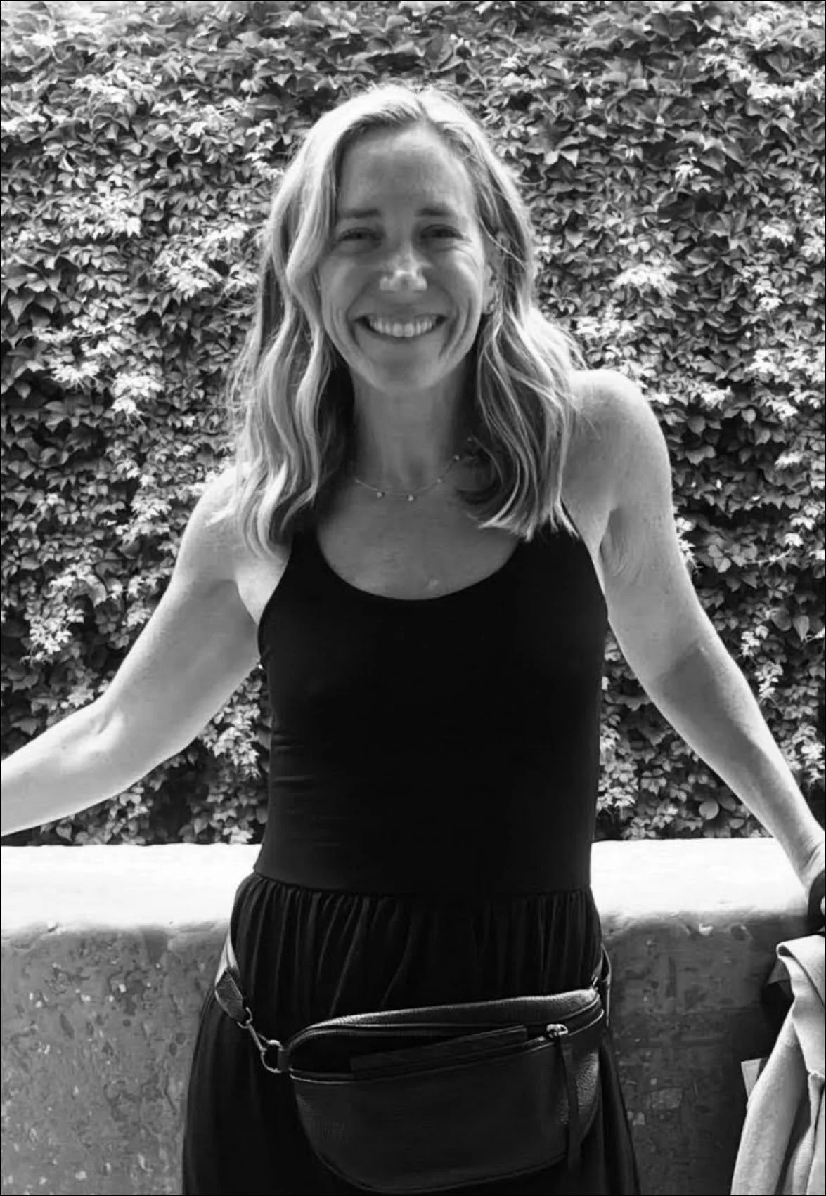A woman with wavy hair smiling, wearing a sleeveless black top and a necklace, standing outdoors in front of a leafy hedge.
