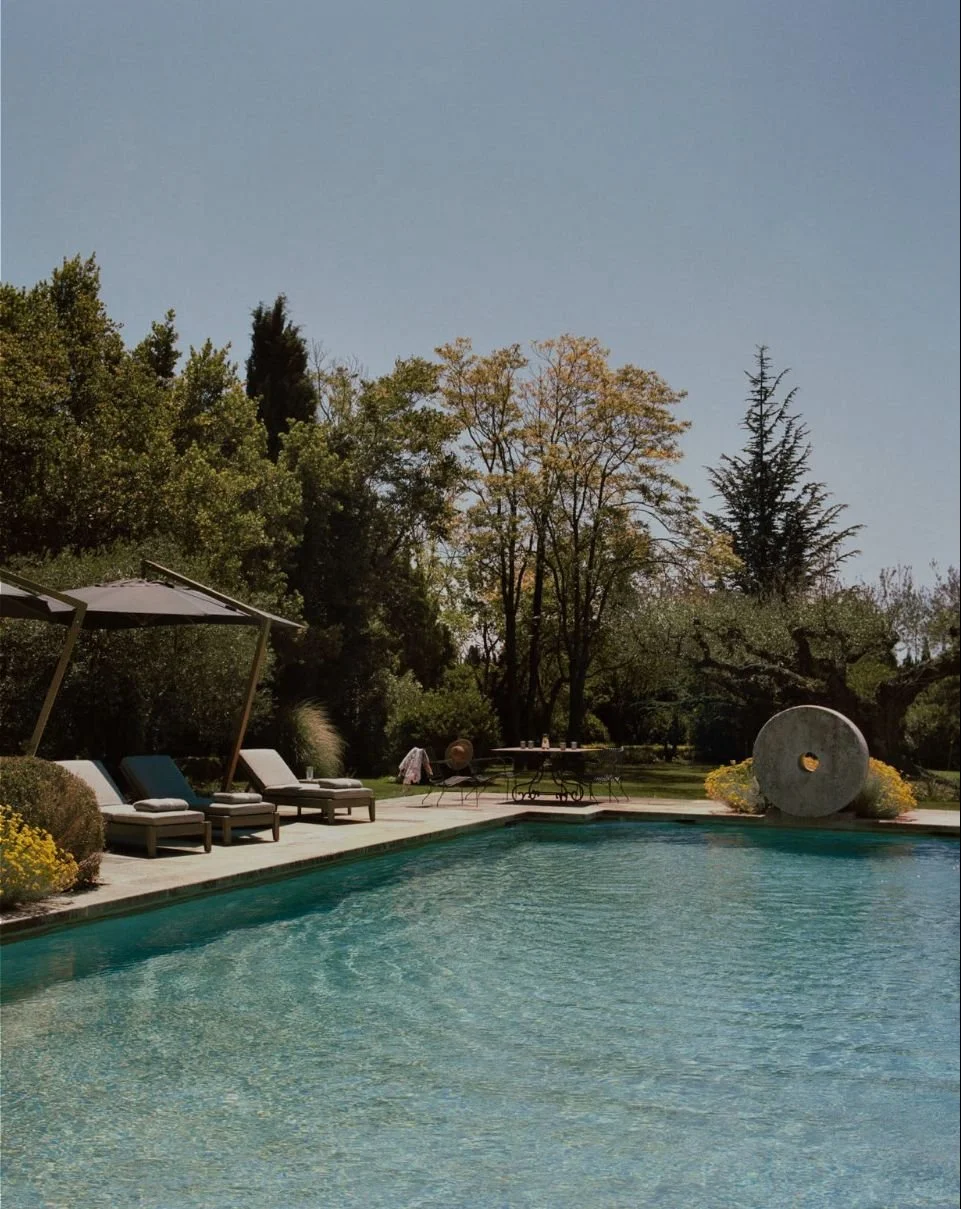 A backyard with a swimming pool, lounge chairs with umbrellas, a round decorative sculpture, and tall trees in the background under a clear blue sky.
