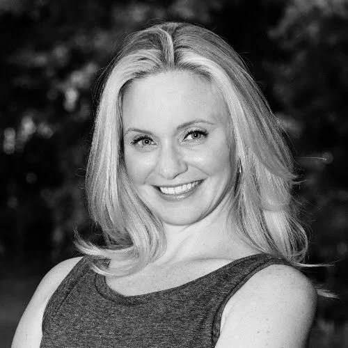 Black and white portrait of a smiling woman with shoulder-length blonde hair, wearing a sleeveless top, outdoors with blurred trees in the background.