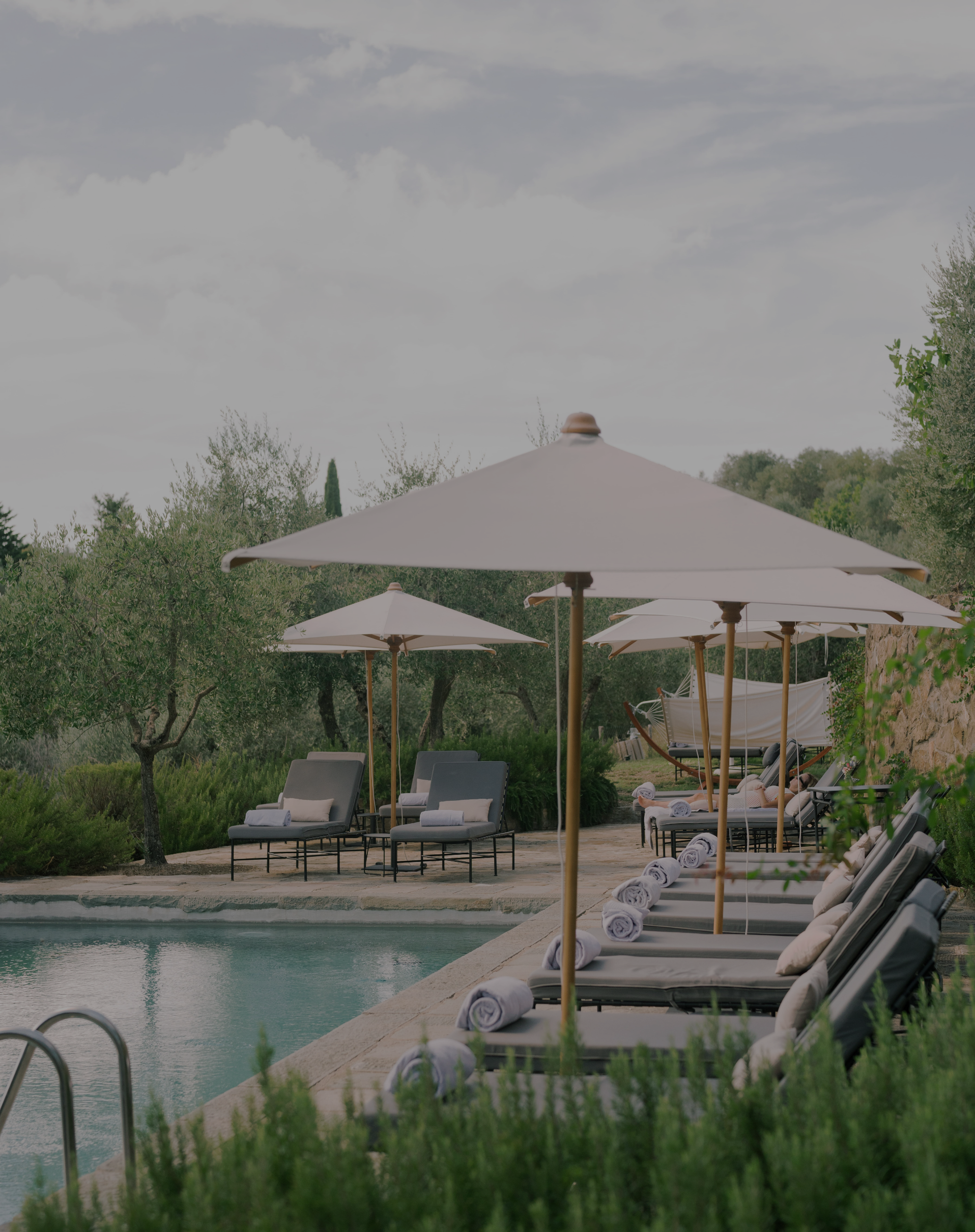 A peaceful outdoor poolside scene with lounge chairs, umbrellas, and rolled towels, surrounded by greenery and trees.
