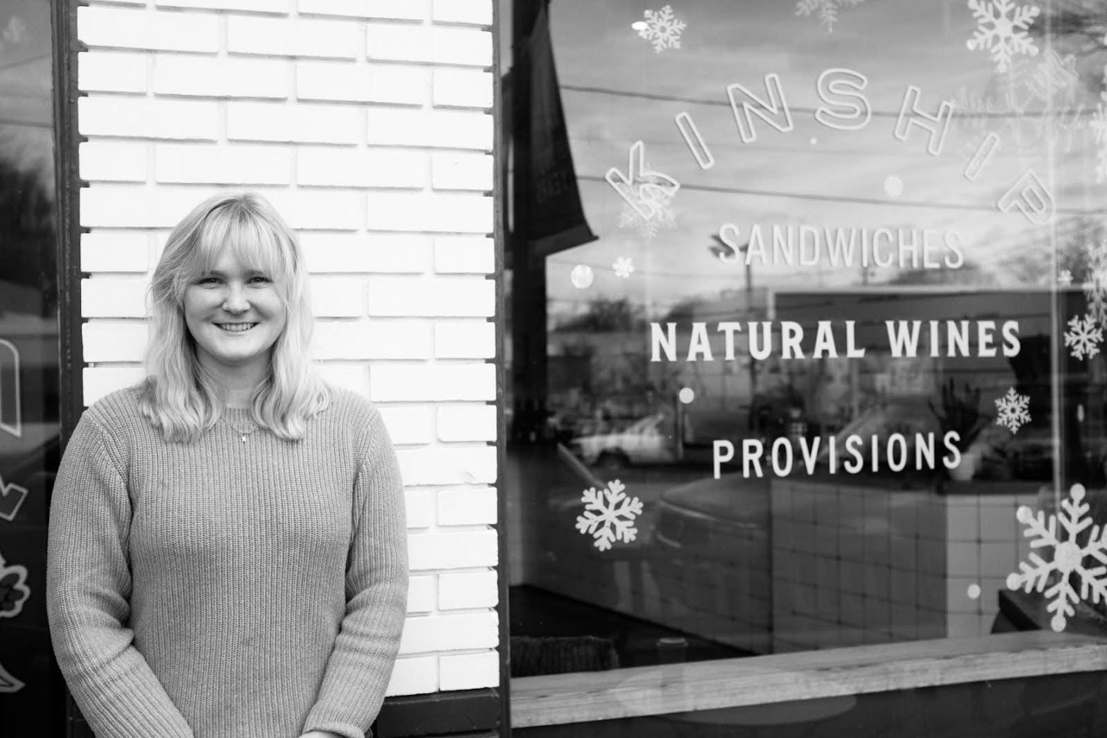 A woman with blonde hair smiling standing outside a shop with holiday decorations and signage for sandwiches, natural wines, and provisions.
