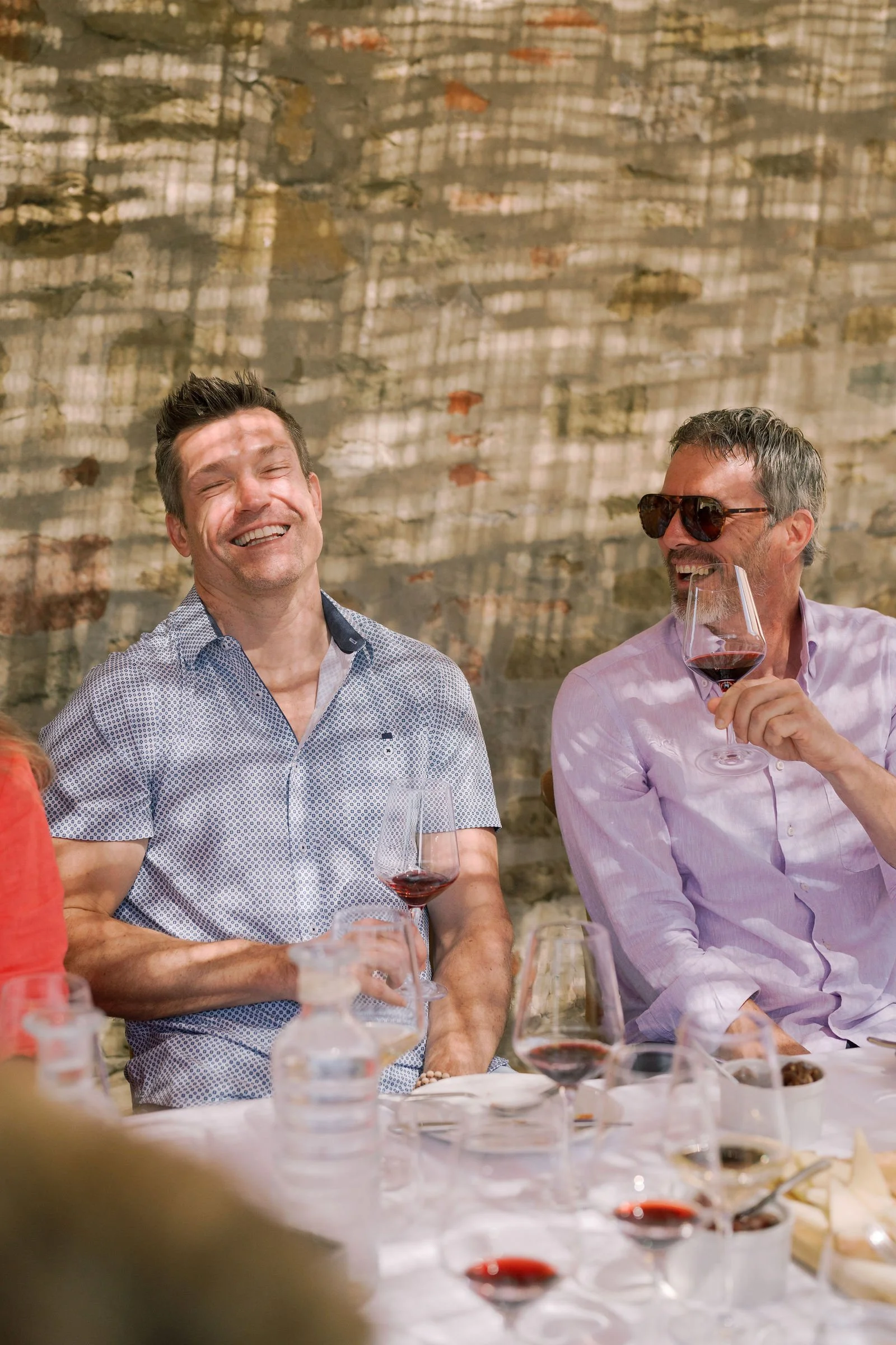 Two men sitting at a dinner table, laughing and enjoying wine, with a brick wall background.