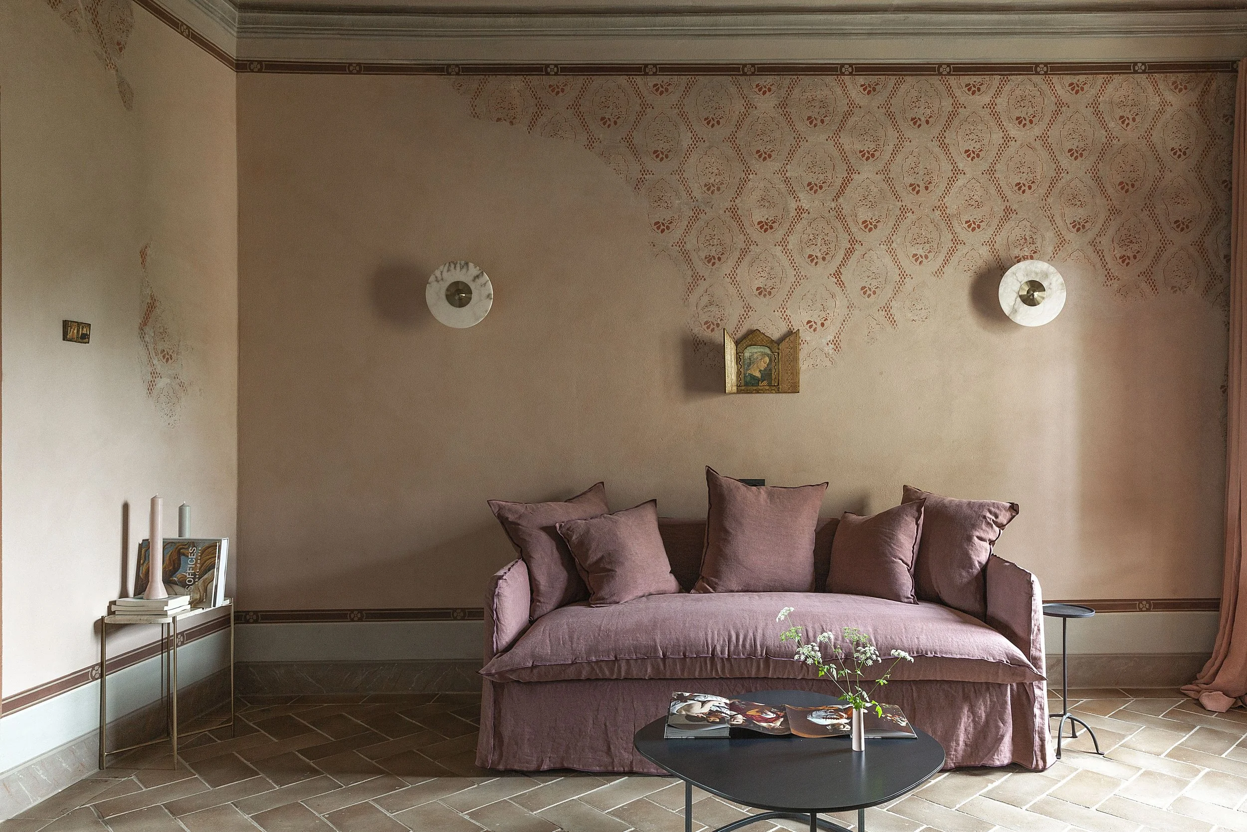 Living room with beige walls, a pink velvet sofa with multiple matching cushions, a black coffee table with a small vase of white flowers and magazines, a side table with books and candles, and a decorative wall hanging above the sofa.