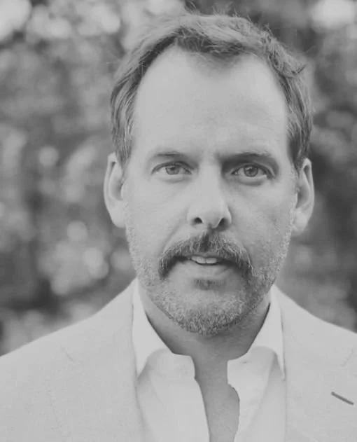 Black and white portrait of a man with short hair, mustache, beard, wearing a light-colored suit jacket and white shirt, standing outdoors with blurred trees in the background.