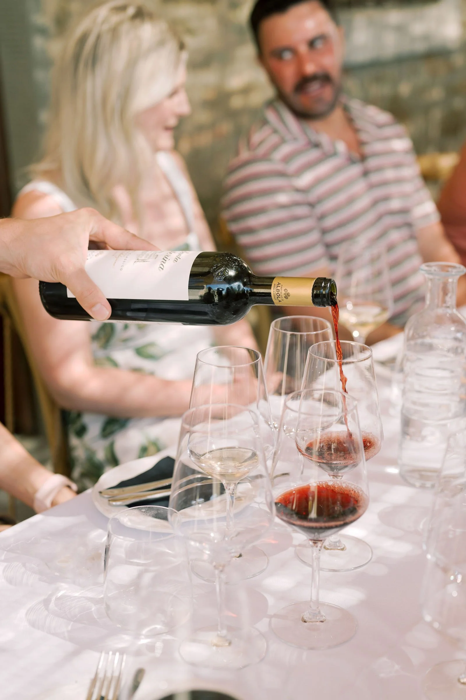 Someone pours red wine into glasses on a table during a social gathering, with two people in the background smiling and talking.