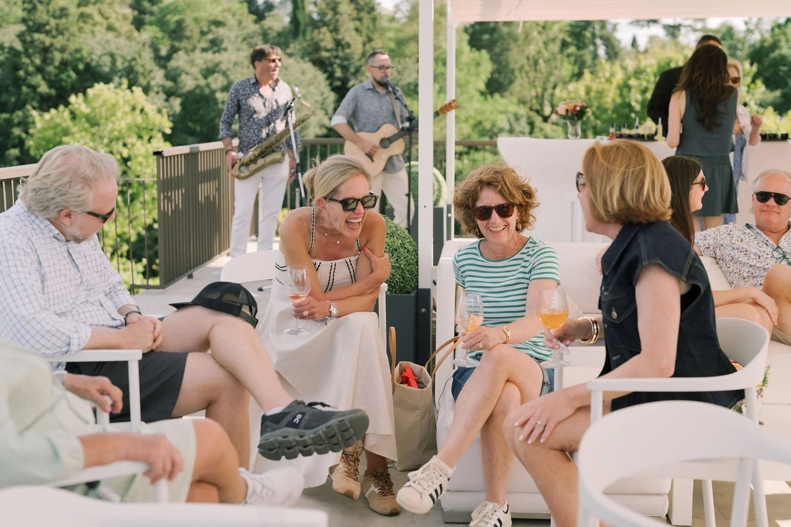 Group of friends enjoying drinks and laughter at an outdoor party with live music in the background.