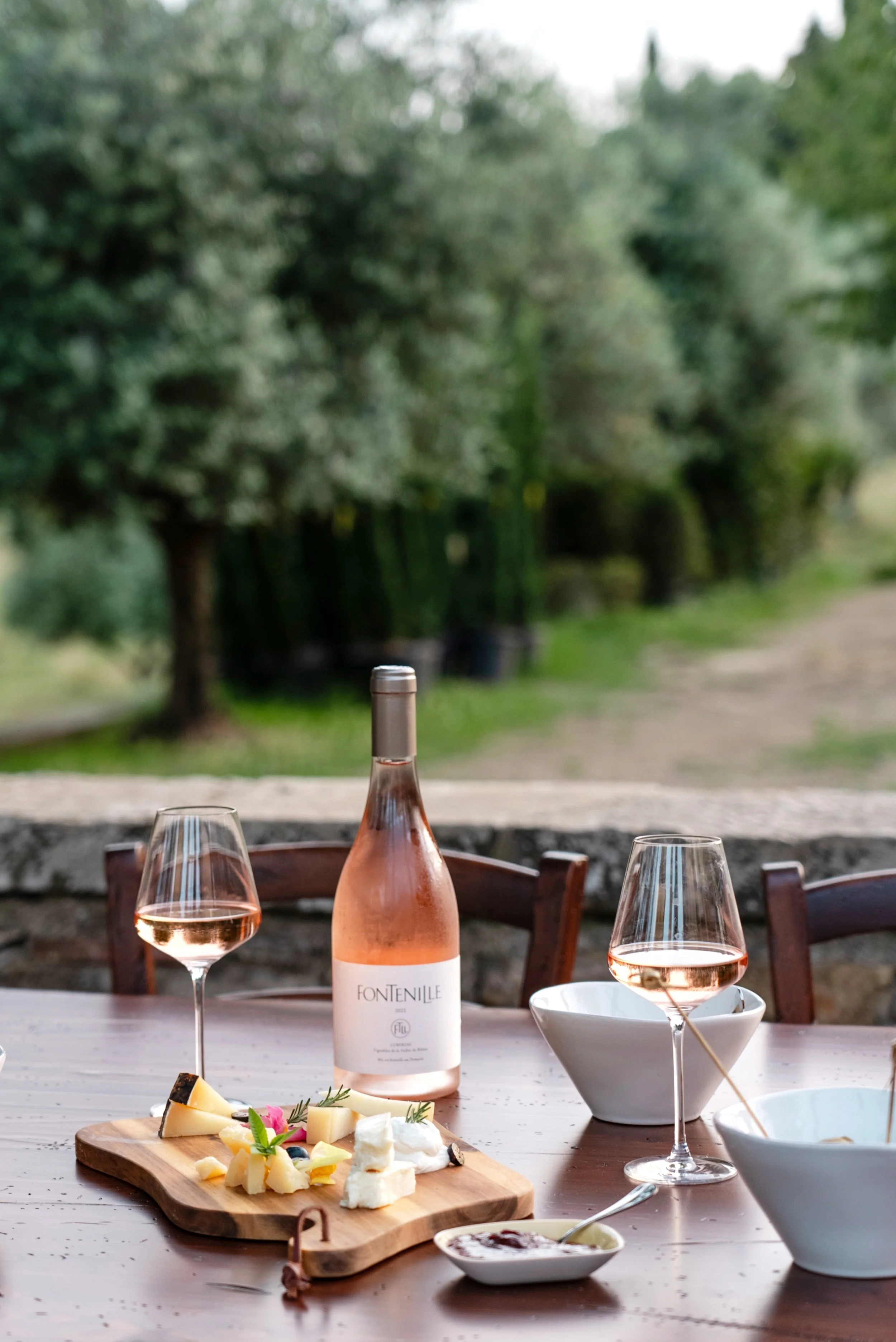 A table set outdoors with a bottle of rosé wine, two glasses of rosé, a cheese platter, and bowls of dip, with a garden in the background.