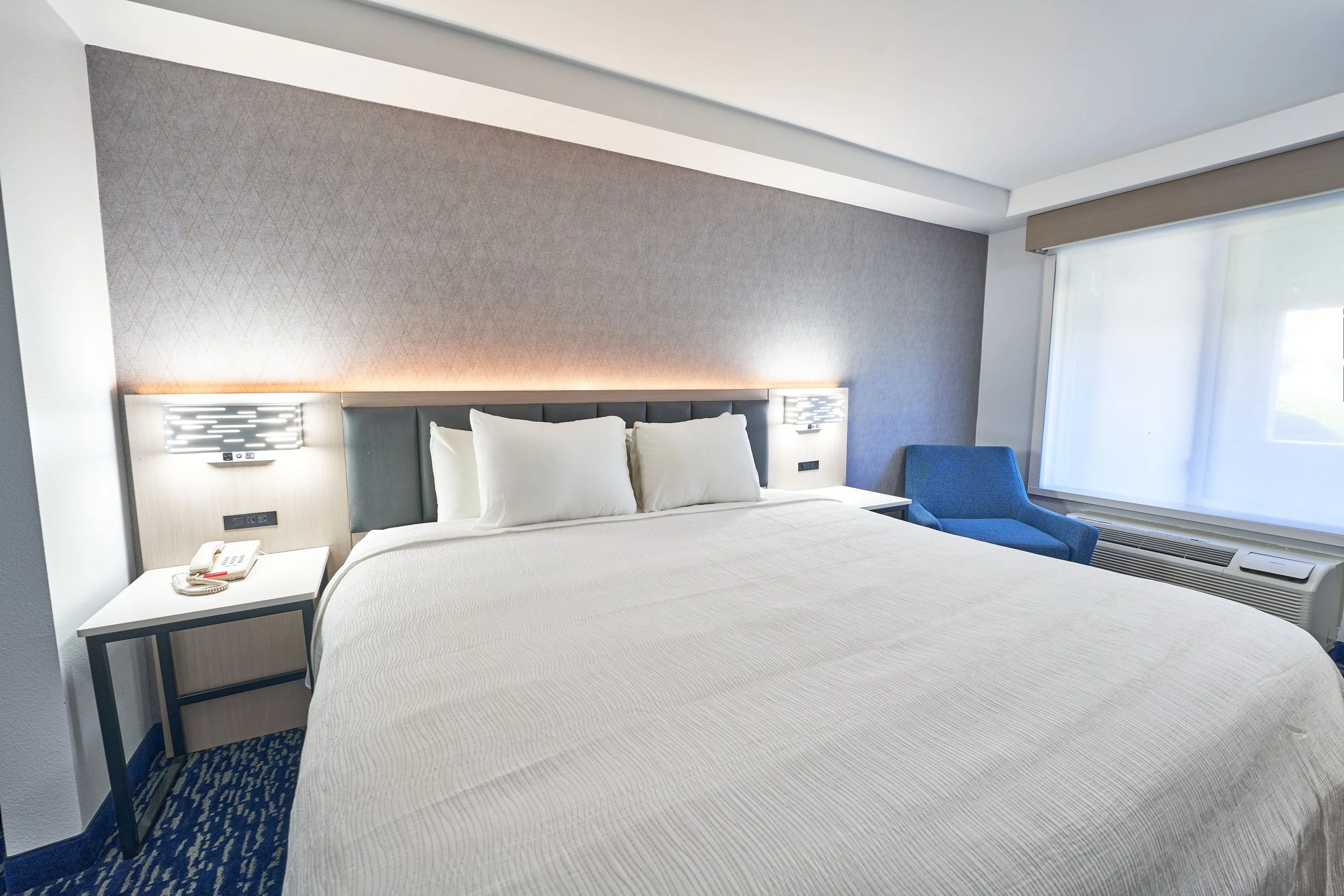 A hotel room with a neatly made bed, white pillows, a blue armchair, a window with white curtains, and a nightstand with a phone.