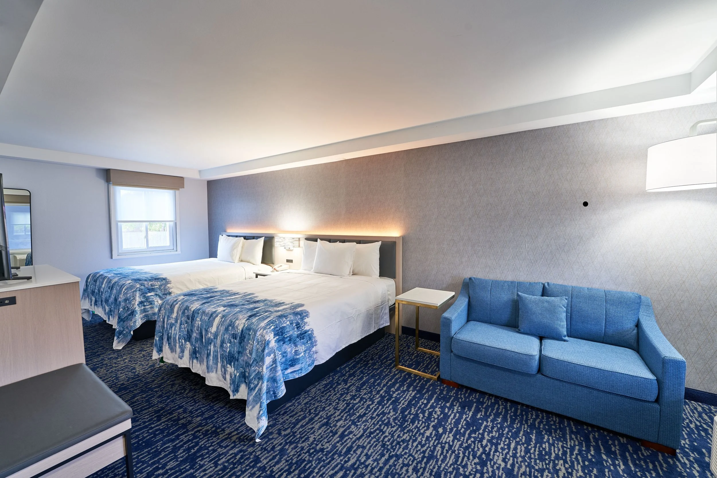 Hotel room with two beds, blue patterned bedding, a blue sofa, a side table, and a window with a view outside.