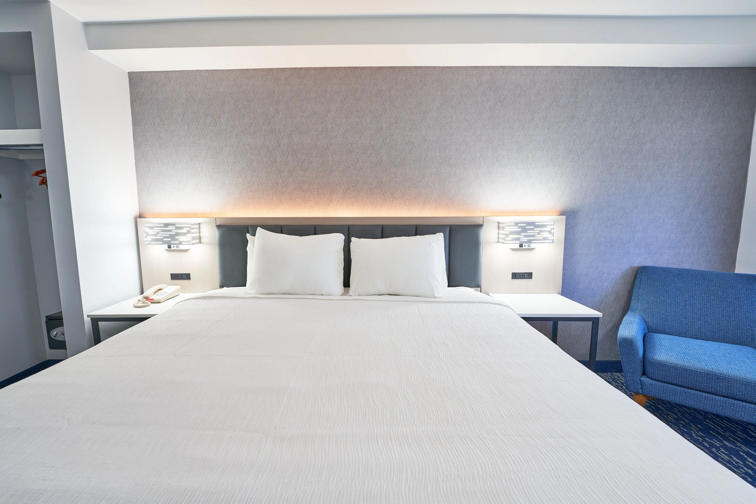 Interior of a hotel room with a large bed, two bedside tables with lamps, a blue armchair, and a built-in closet.