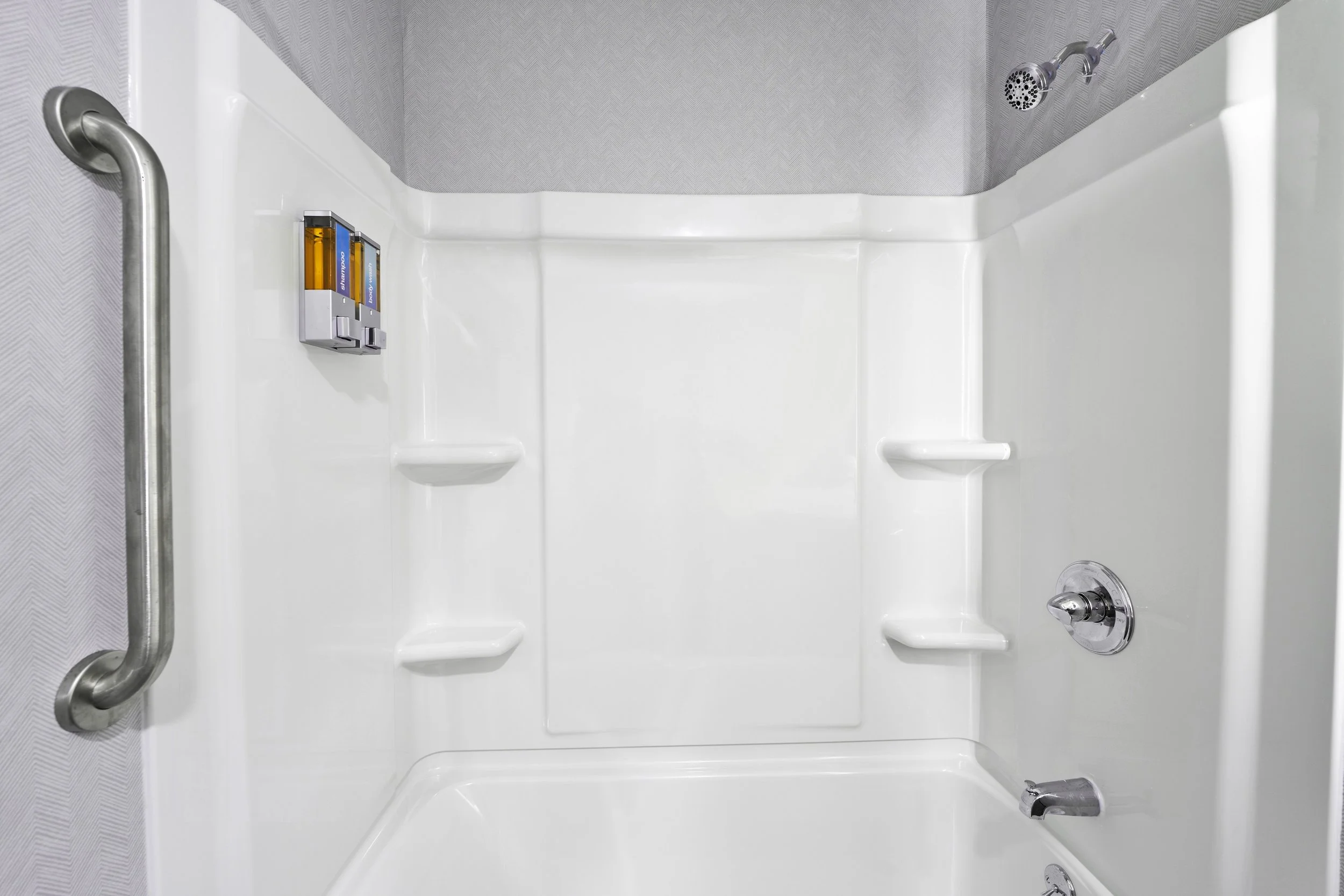 Empty white shower stall with a silver grab bar on the left, soap holders built into the walls, and a showerhead on the top right.