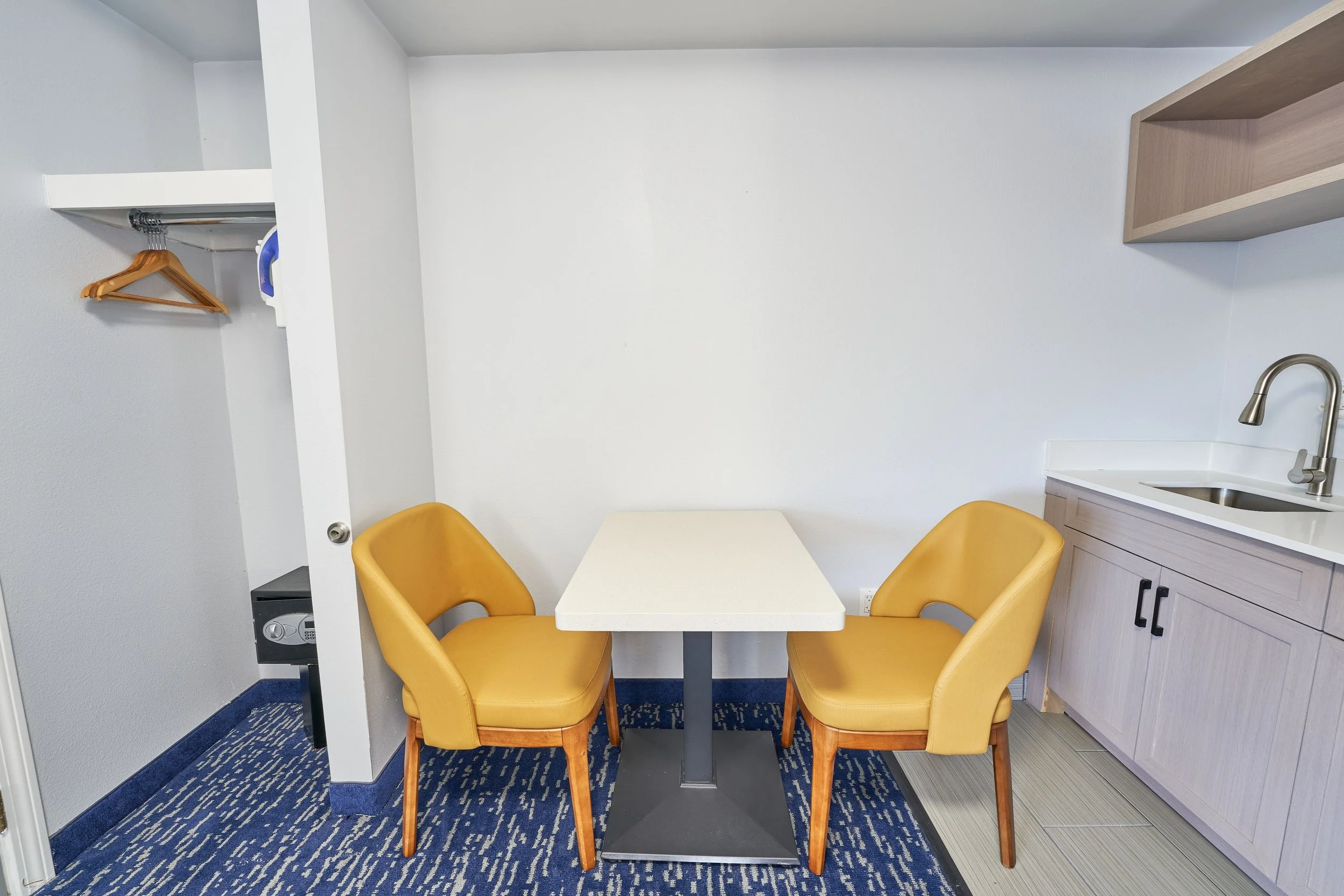 Small dining area with two yellow chairs, a white table, a kitchenette with a sink and light wood cabinets, a small closet with hangers, and a patterned blue carpet.