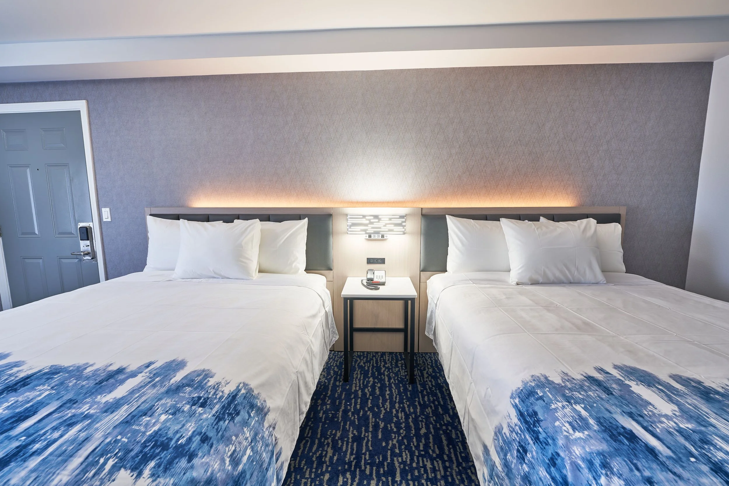 Two large beds with white bedding and pillows in a hotel room, separated by a nightstand with a telephone and remote control, against a textured gray wall with soft lighting.