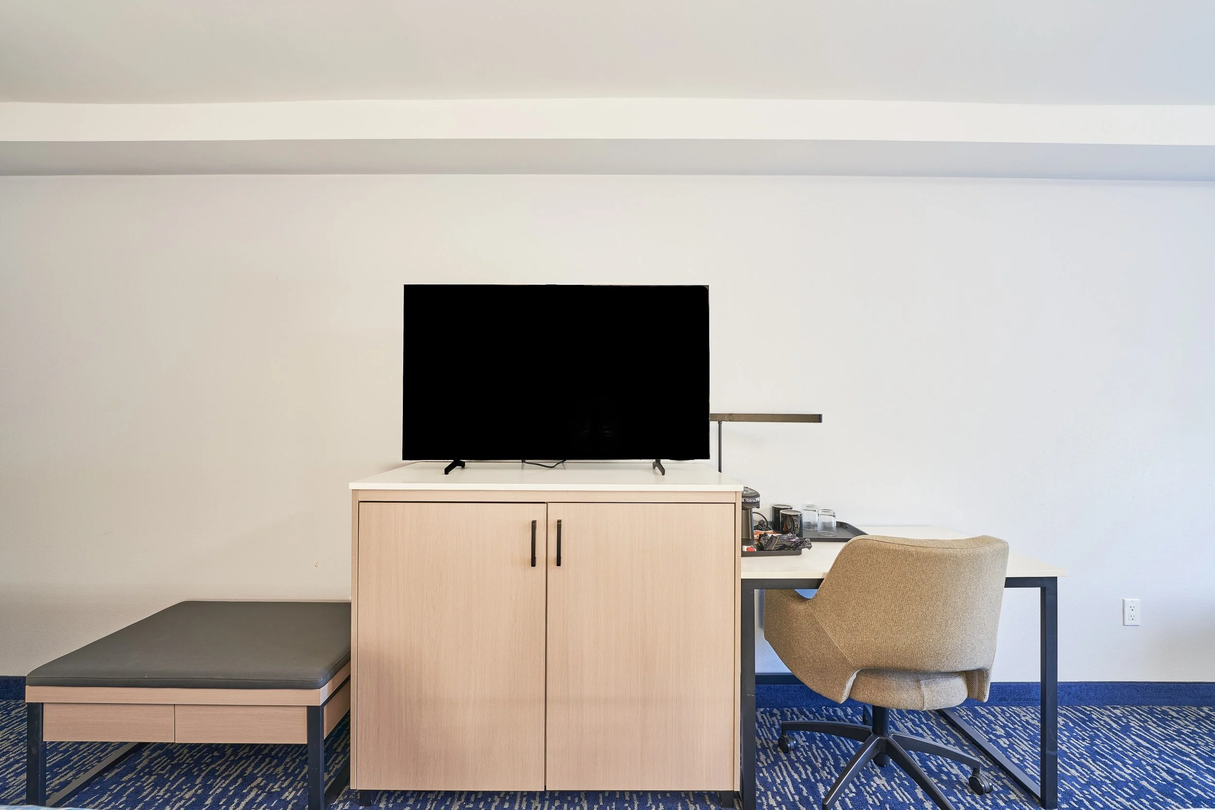 An empty hotel room with a flat-screen TV on a cabinet, a desk with a beige chair, and a bench, against a plain white wall and a blue patterned carpet.