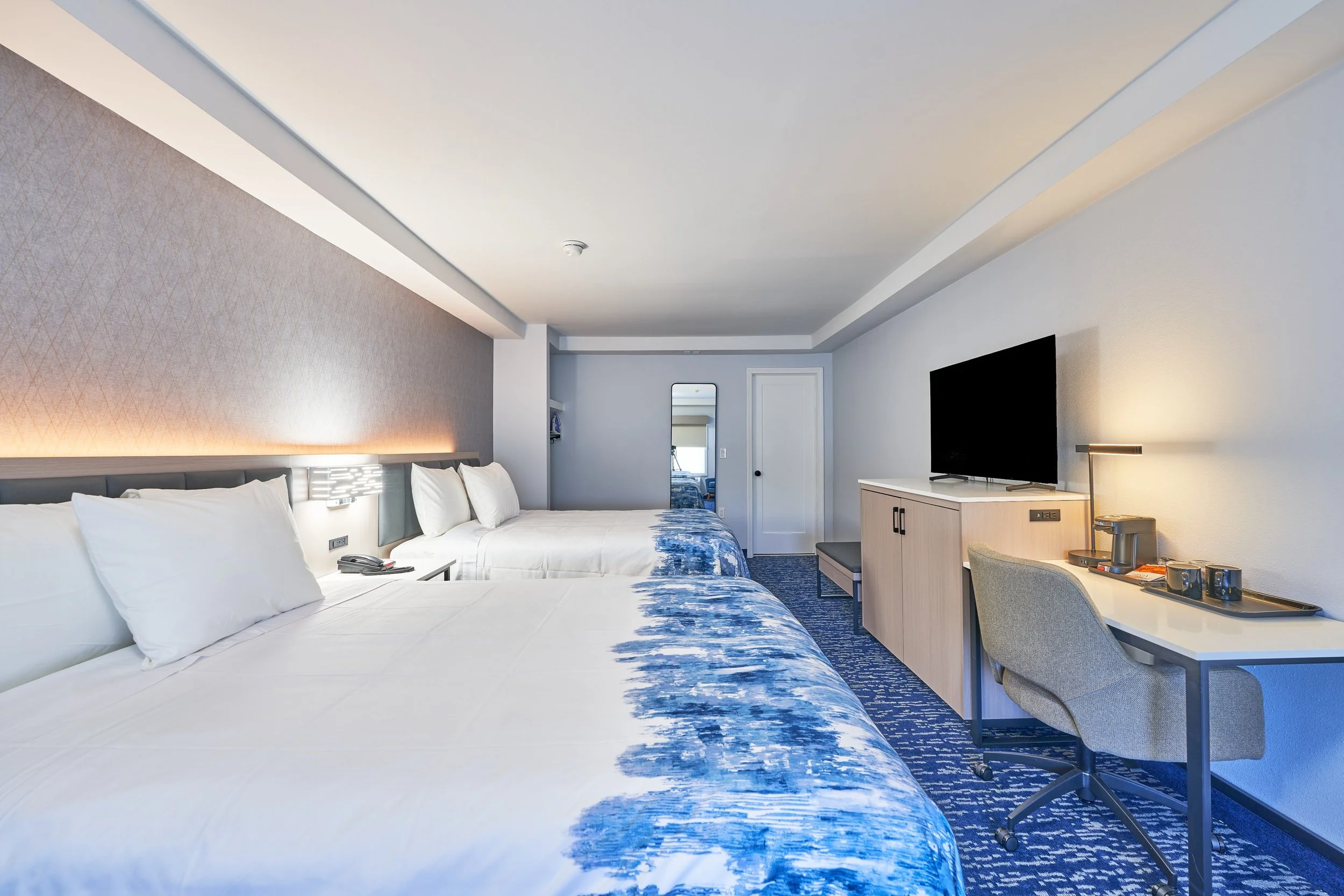 Hotel room with two beds, a TV, desk, and mirror, decorated in neutral and blue tones.