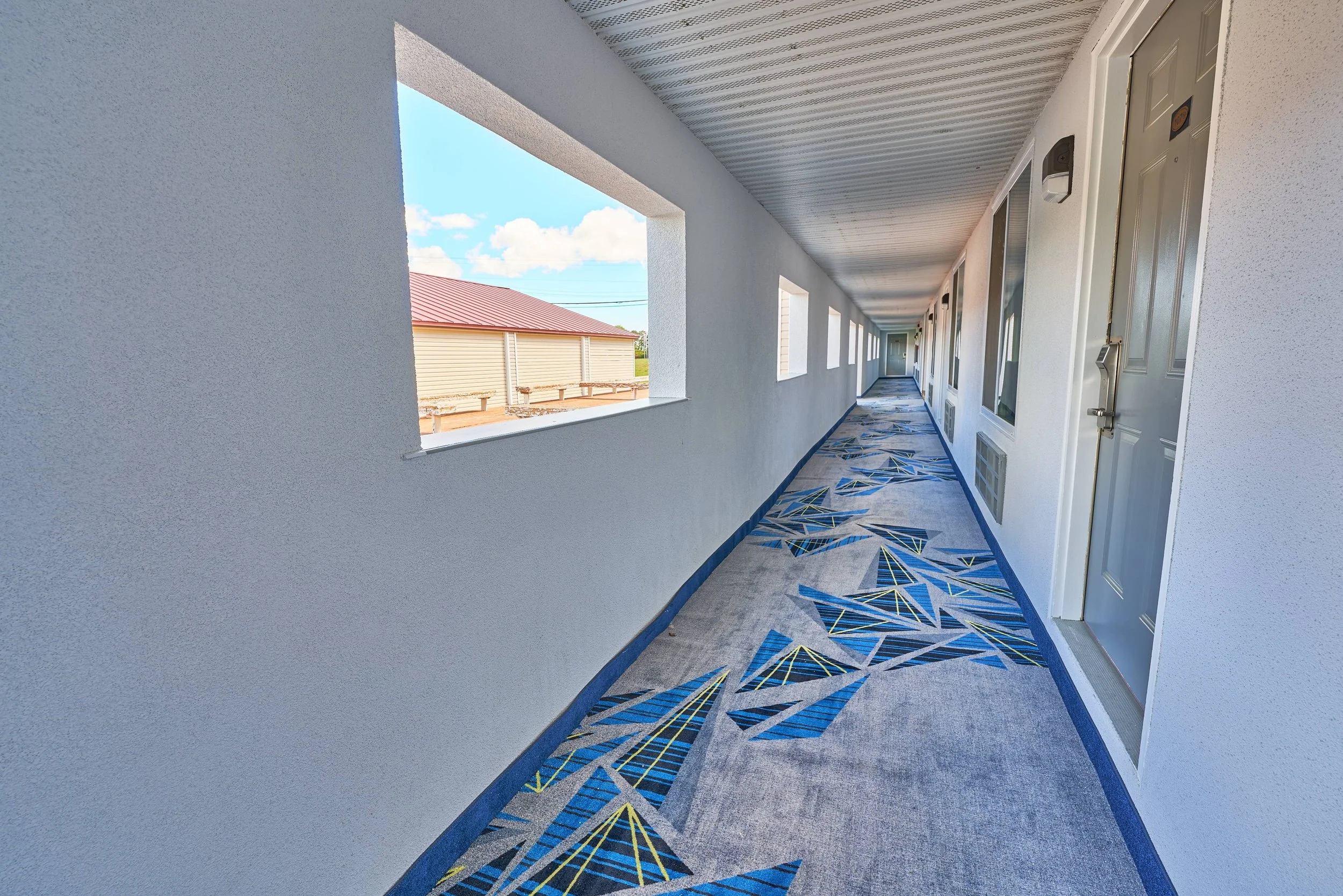 Long outdoor balcony corridor with blue and gray patterned carpet, white walls, and multiple apartment doors on the right. Large rectangular open windows with a view of a neighboring building with a red roof on the left, and a blue sky with clouds in