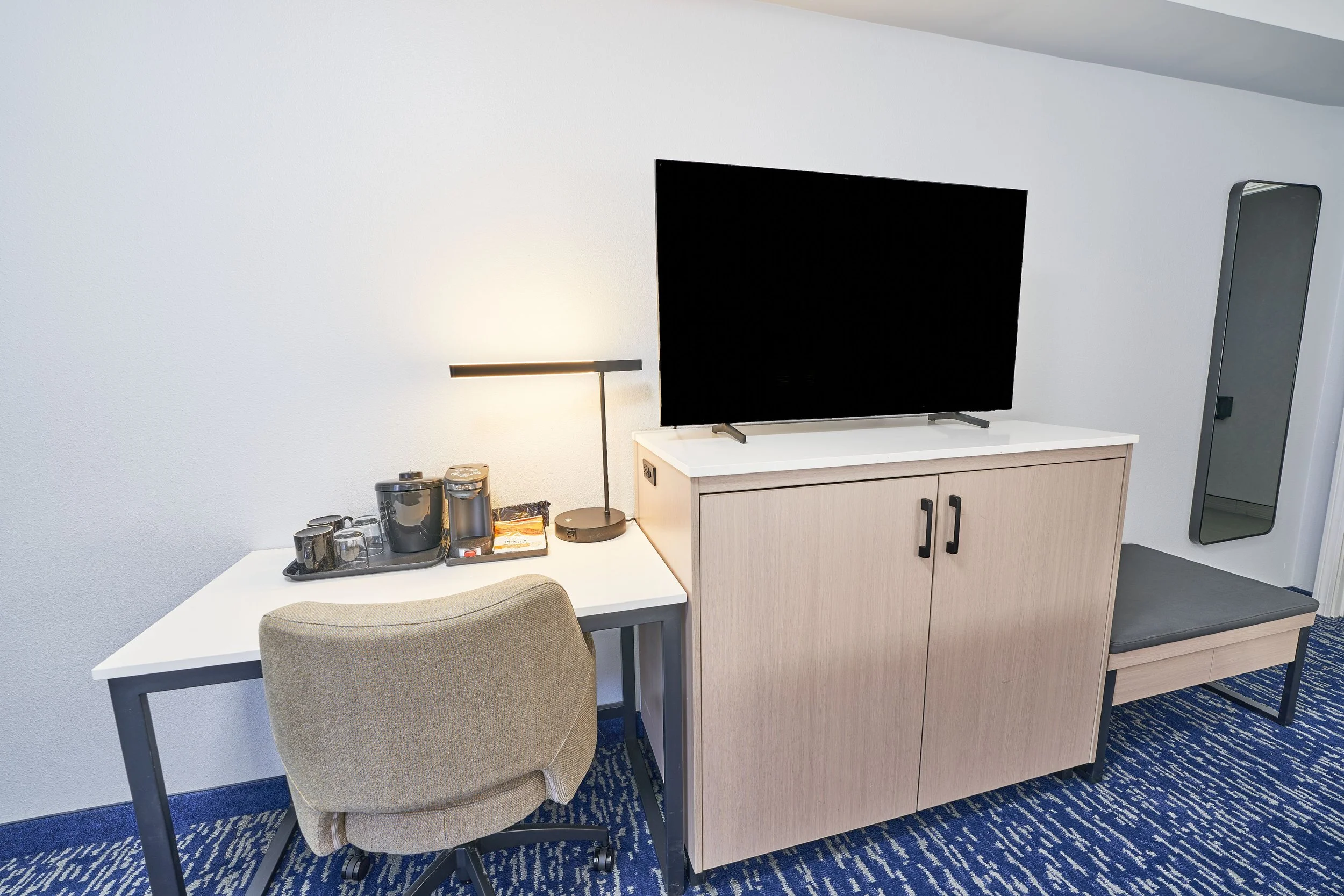 Hotel room workspace with a desk, chair, coffee set, lamp, television on a cabinet, bench, and mirror, against white walls and blue patterned carpet.