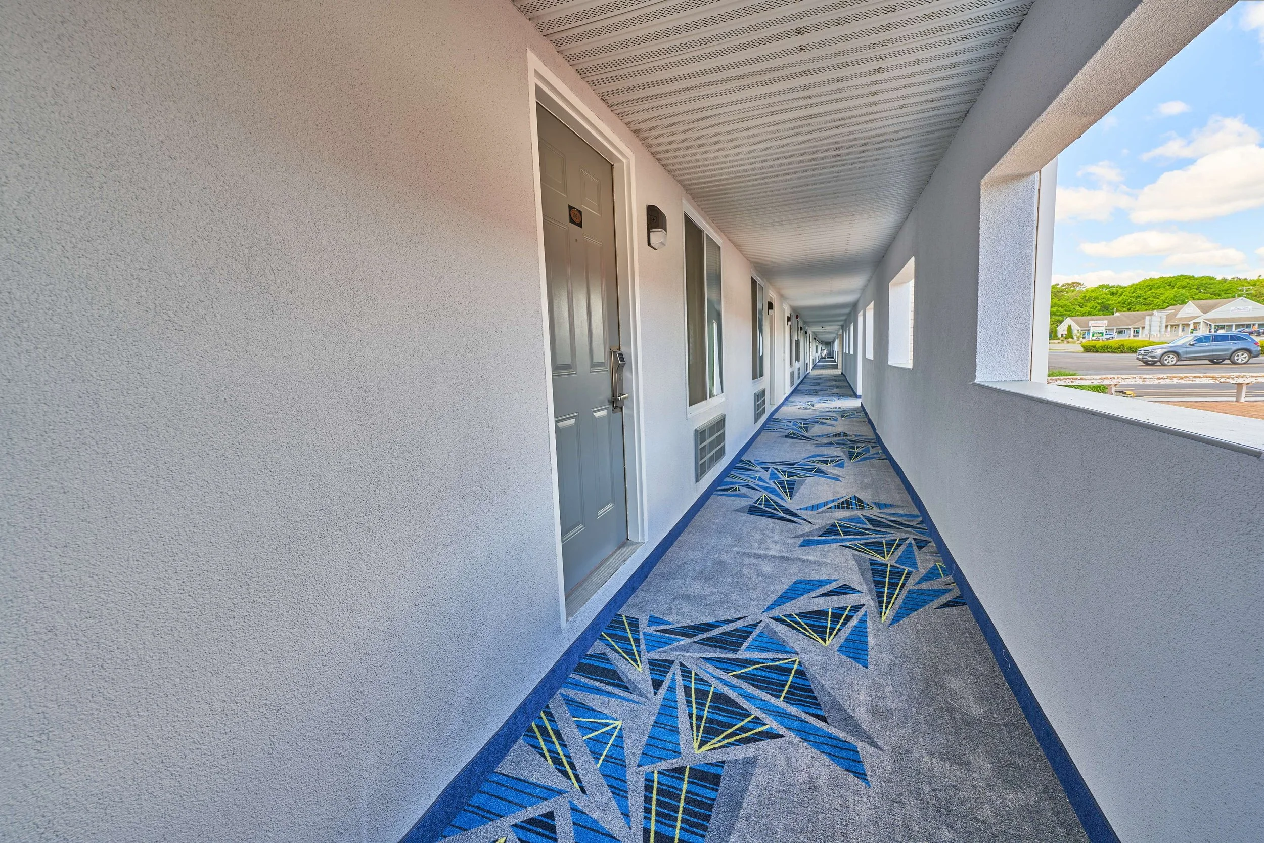 Long outdoor corridor with white walls, gray doors, colorful geometric patterned carpet, and open windows showing a parking lot and green hills in the background.
