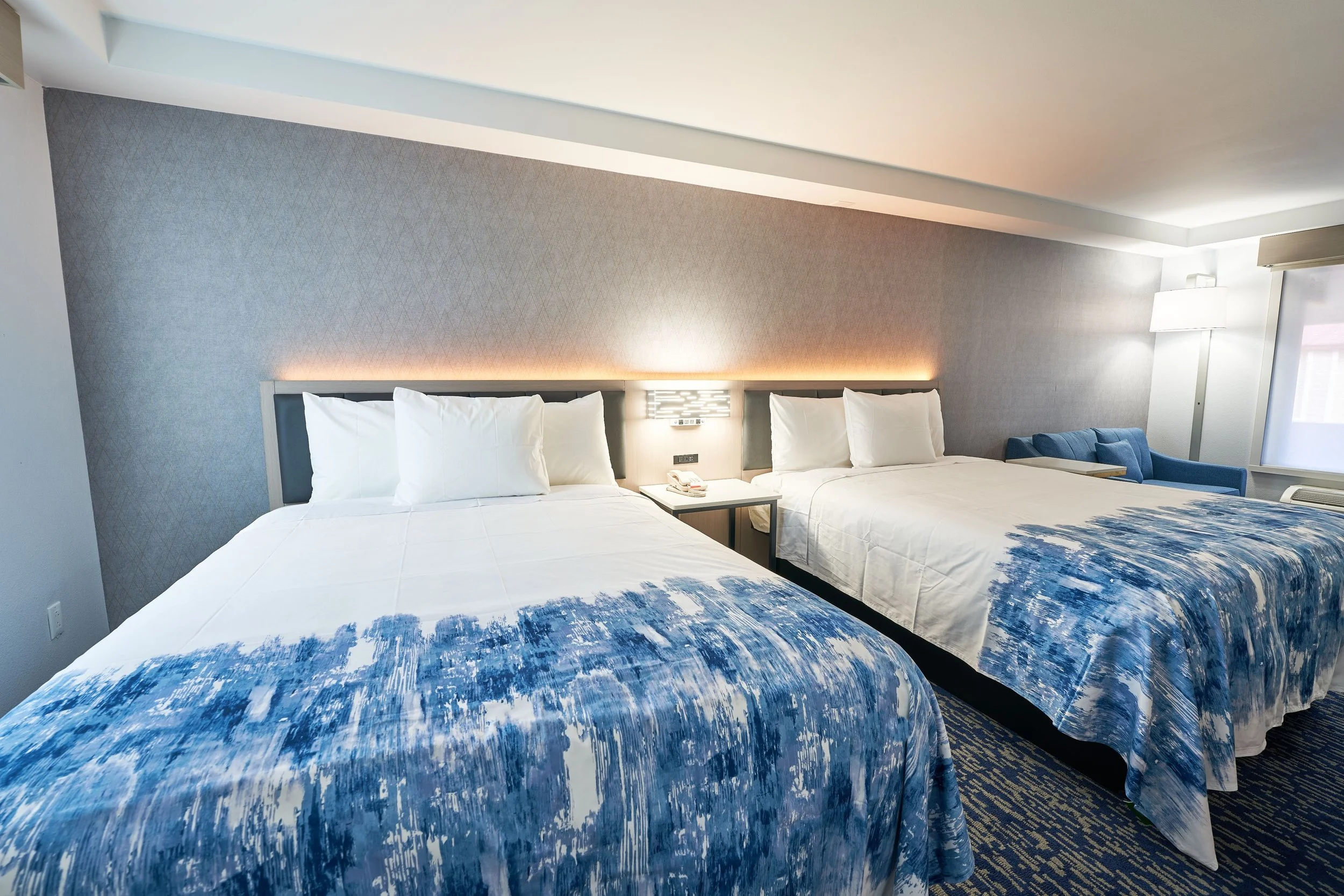 Two double beds in a hotel room with white bedding, blue patterned bed runners, and pillows. A nightstand with a landline phone sits between the beds, and a blue armchair is near the window on the right side.