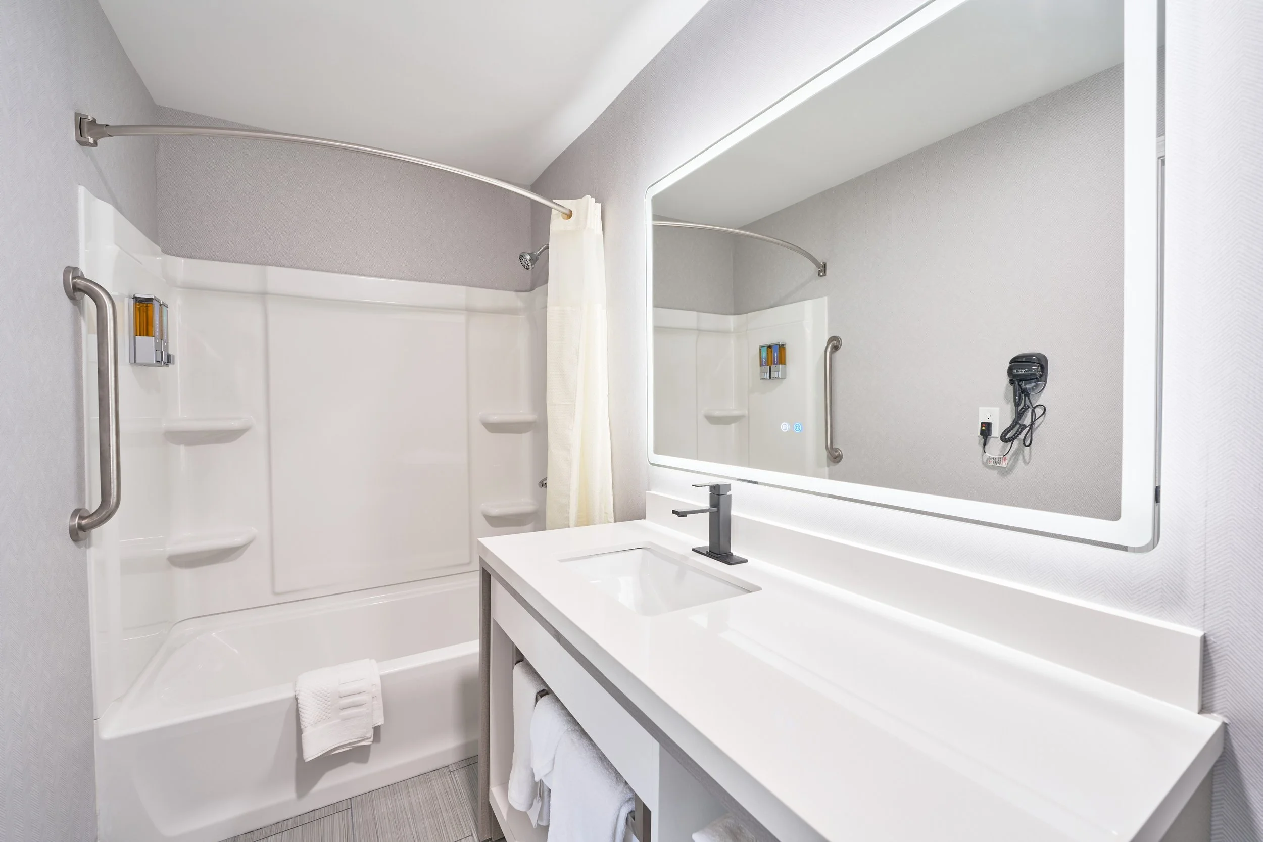 Modern bathroom with a white vanity, large mirror, and a shower area with a curved curtain rod and grab bars.