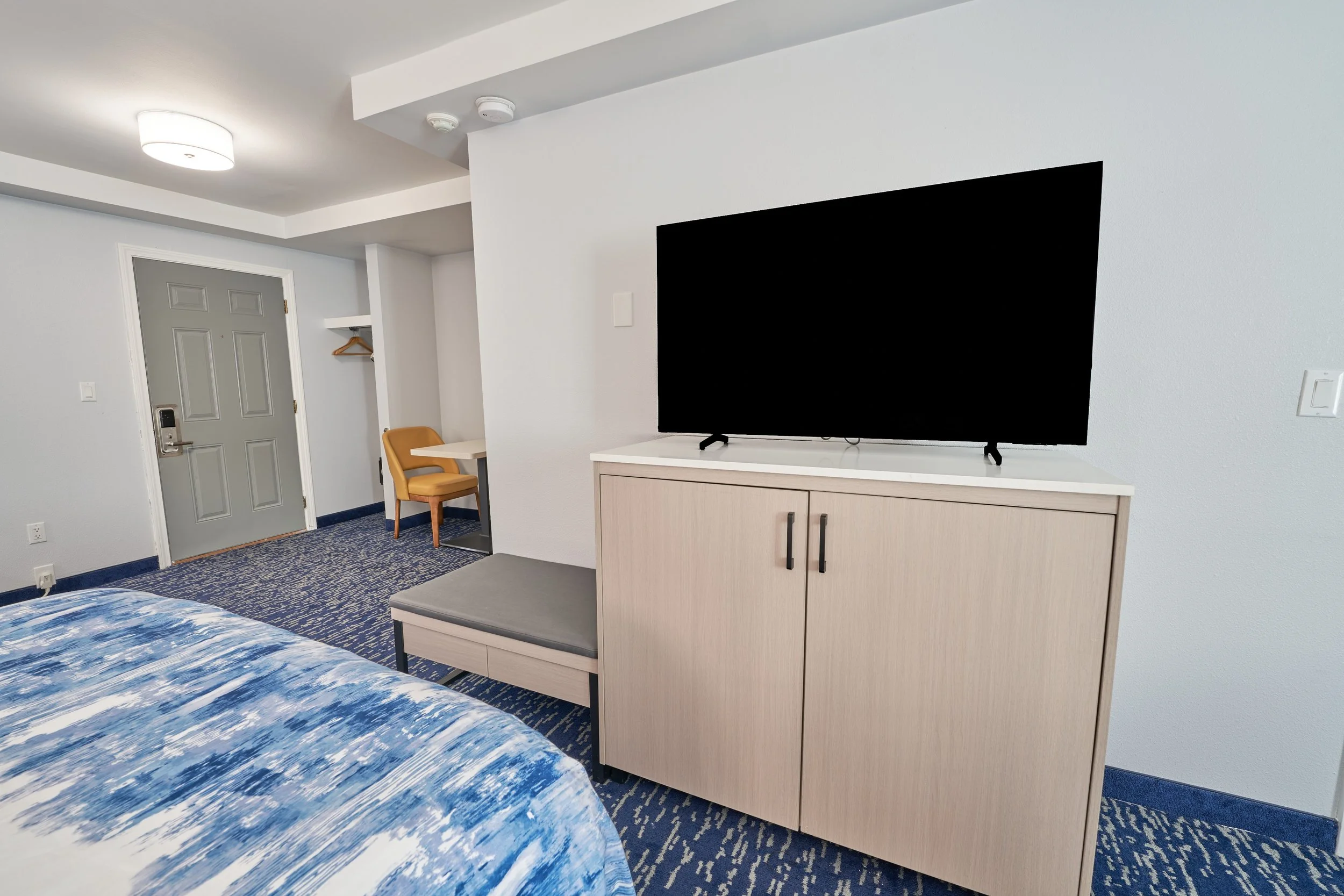 Hotel room with a wall-mounted TV on a light-colored cabinet, a bed with a blue and white duvet, and a small seating area near the door.