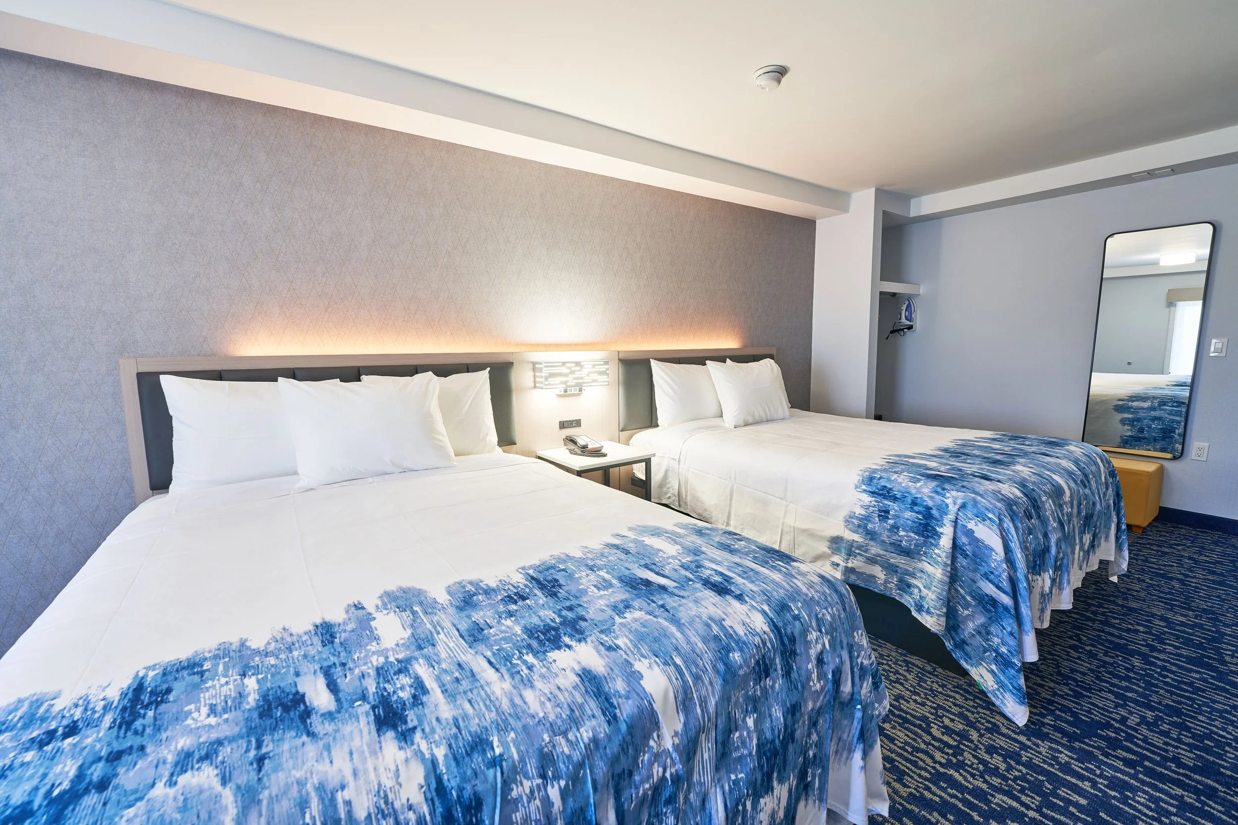 Two double beds with white linens and blue patterned bedspreads in a hotel room, with a wall-mounted headboard, a small nightstand, and a large mirror on the wall.